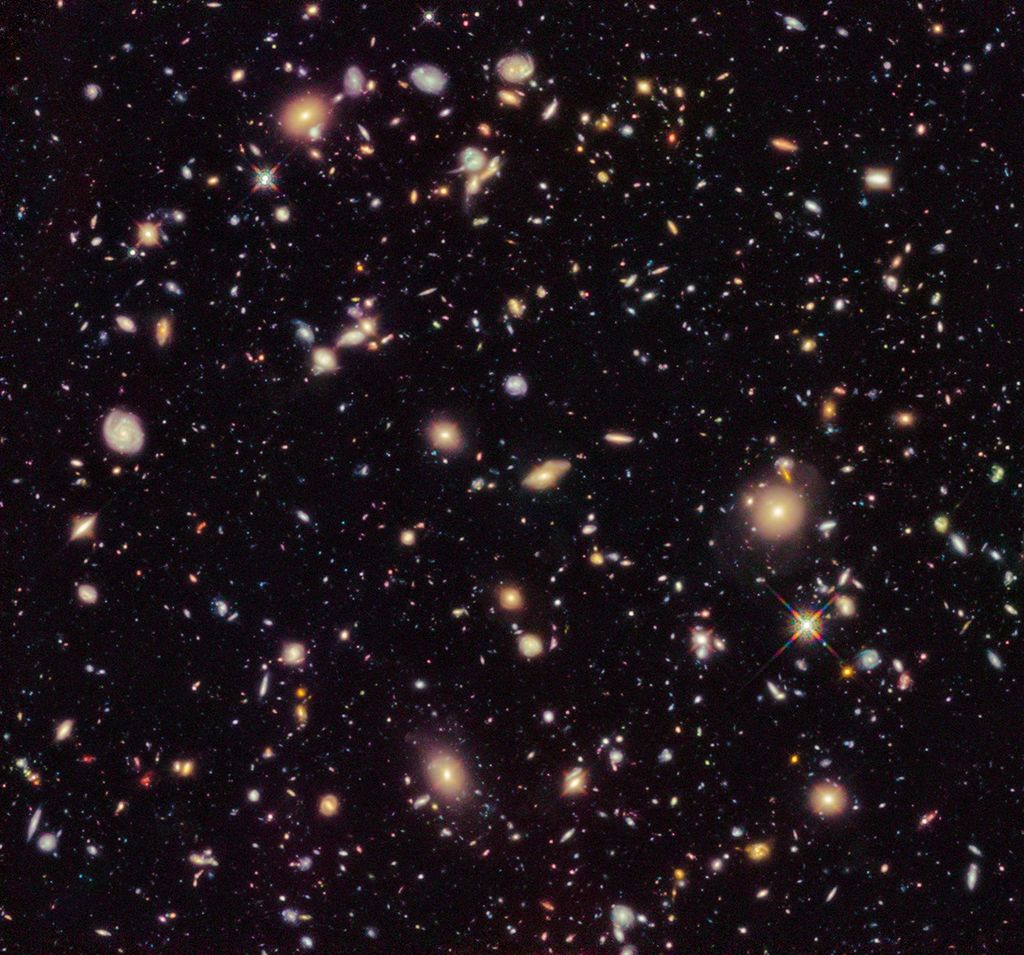Image is filled with distant galaxies in colors of yellow-gold, red, blue, white. The whole image has a reddish tint.