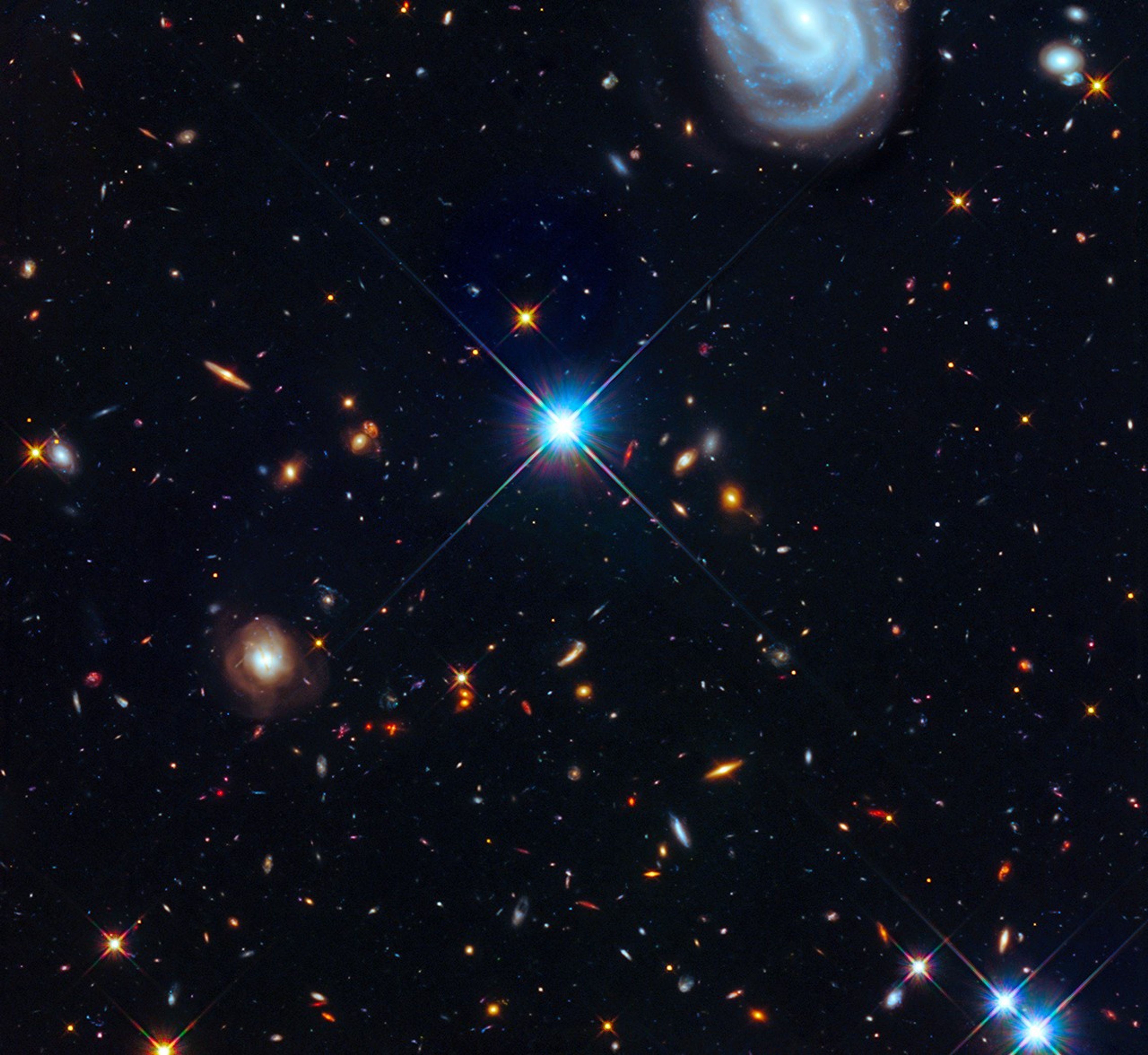 Hundreds of galaxies in a diverse array of shapes and colors are scattered across the dark background of space. Bright stars are visible near the center and in the bottom-right. A large blue-white spiral galaxy can be seen at the top on the right side.
