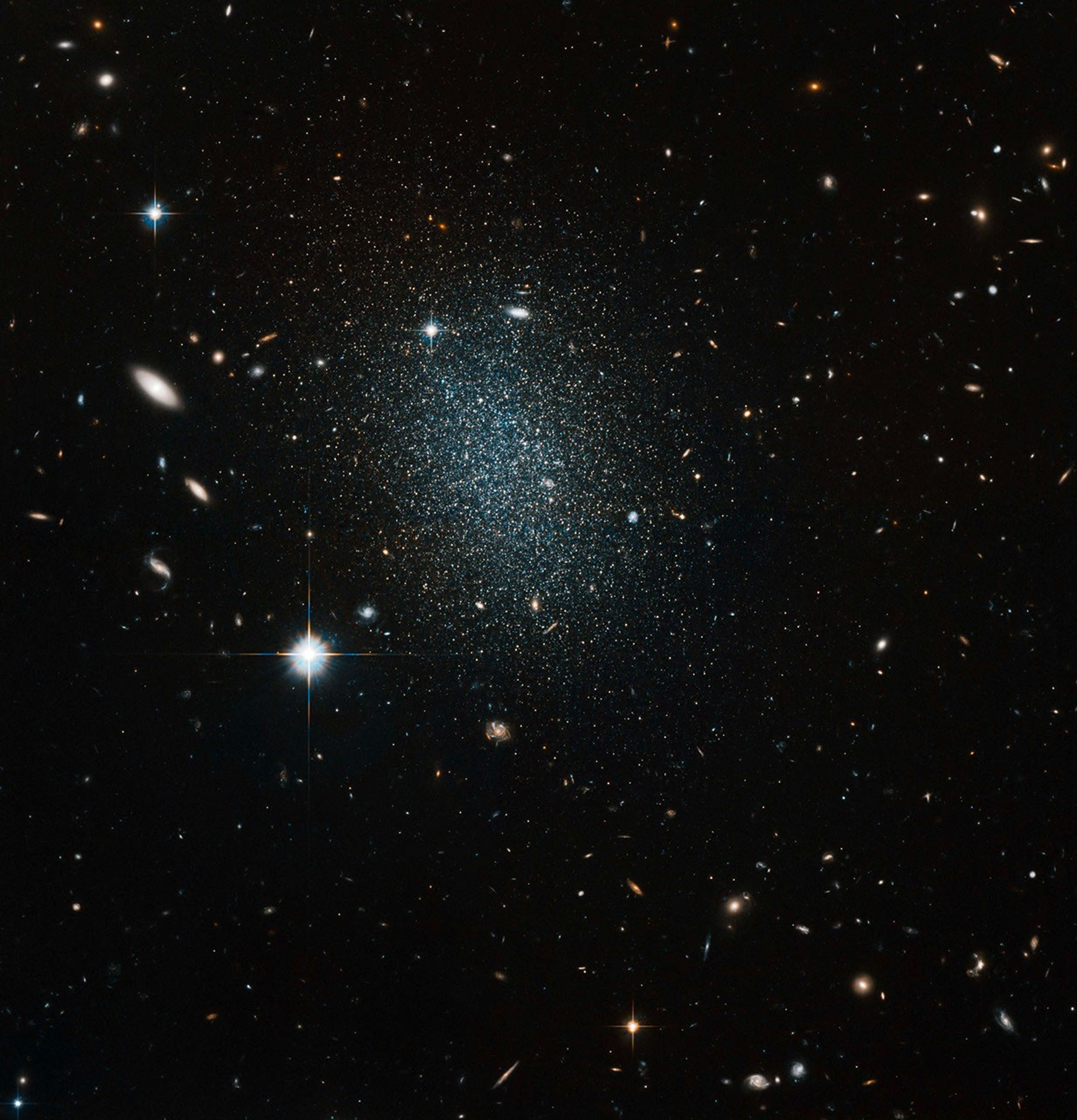 A black background field full of galaxies and stars, with one bright foreground star left of center. Just above-left of center, the galaxy appears as a fuzzy patch of stars.