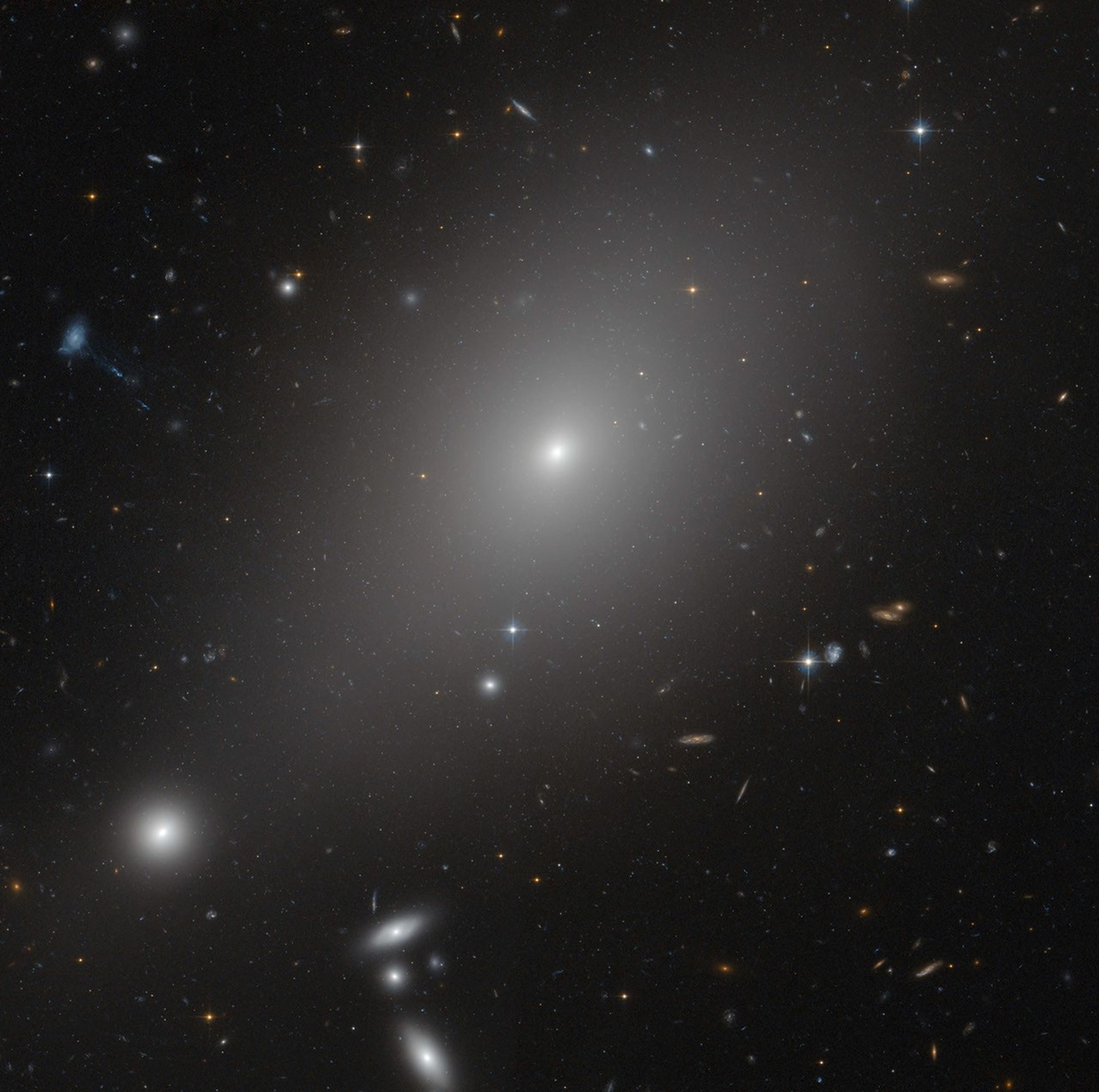 A hazy white light shines just above the center of the image, against black space dotted with faint, distant stars and galaxies. Several more white galaxies glow distantly in the bottom-left.