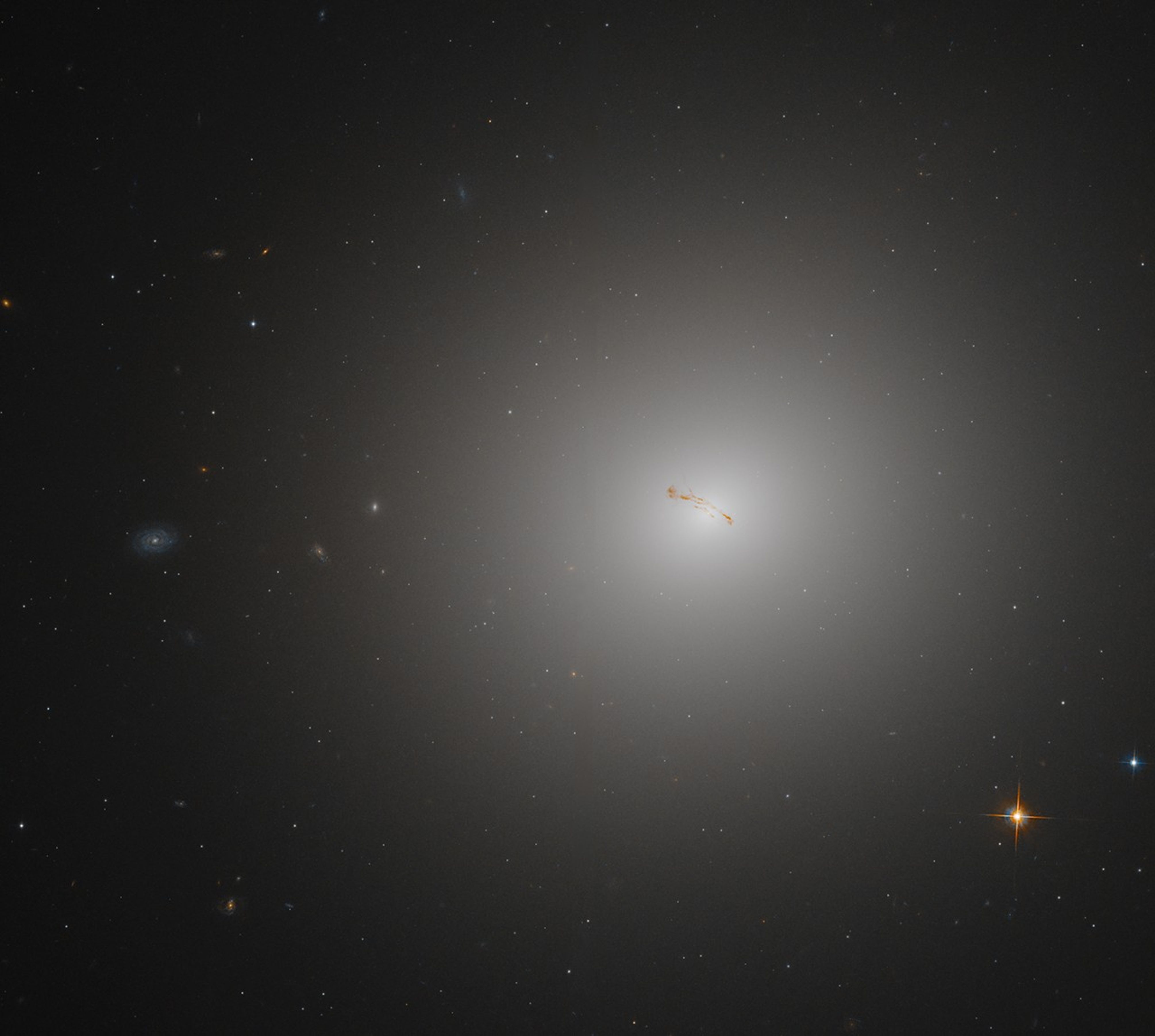 M84 fills most of the right side of the image. The galaxy appears as a bright,-white, hazy ball against a black background dotted with other galaxies. The galaxy's core is bright-white, transitioning to hazy, diffuse white toward the galaxy's outer edges. Bands of rusty-red dust stretch across the galaxy's core.