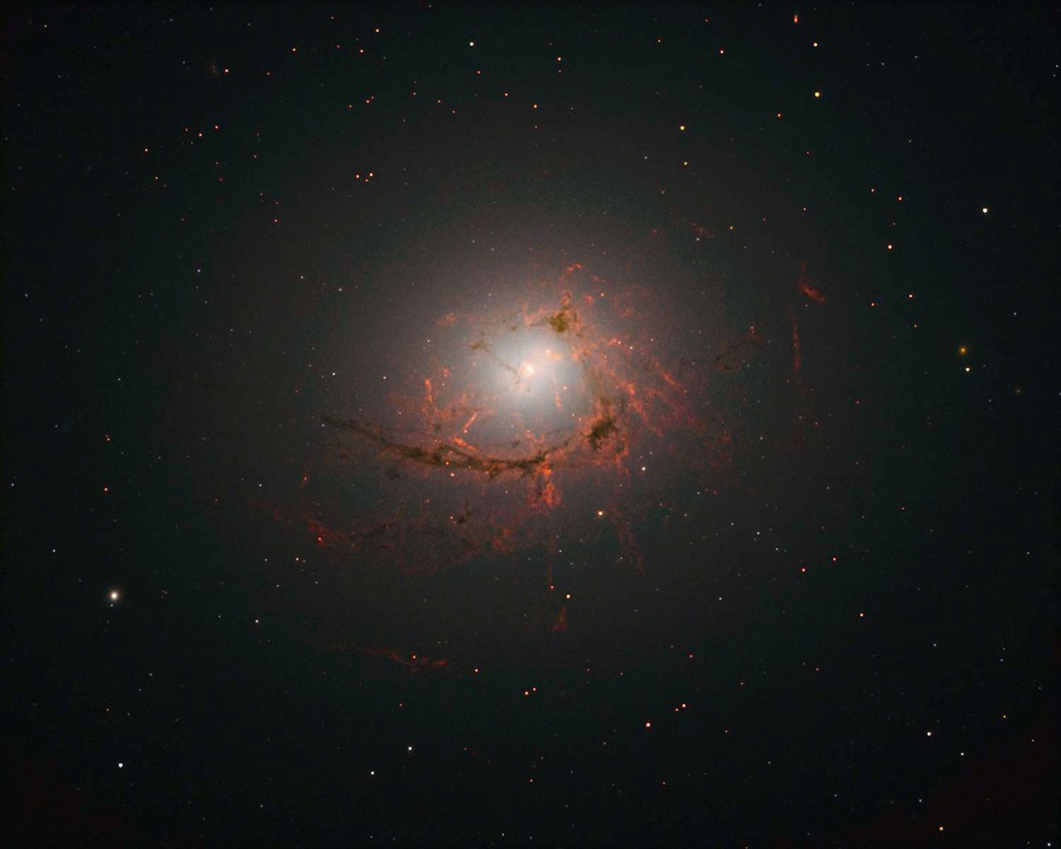 Red and black filaments of gas and dust surround a glowing white core, resembling a ring of flames. Hundreds of other stars and galaxies dot the black background.