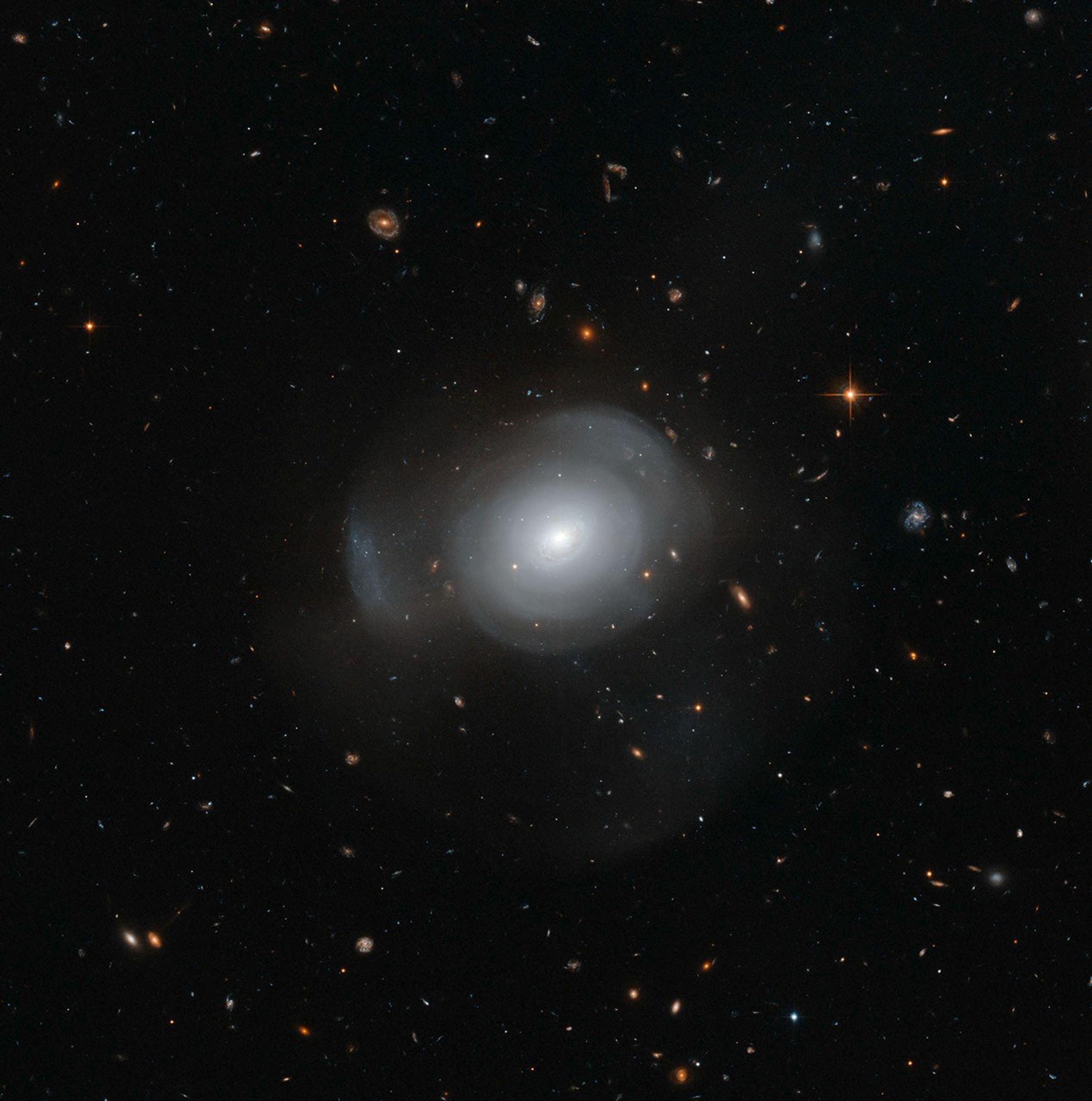 Bright, white diffuse sphere with an extremely bright core. Diffuse shells of matter form layers around the core. Hundreds more galaxies of different shapes and colors shine in the background.
