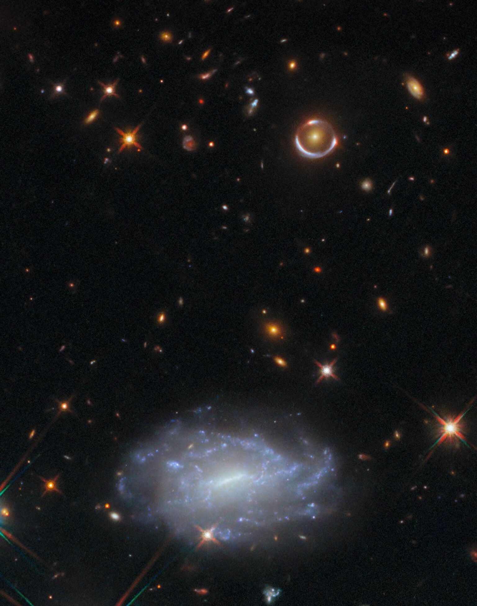 Many mostly small, bright objects scattered over a dark background in space. In the top half on the right is an elliptical galaxy, a round light larger than the others, with a slightly warped ring of light around it. In the bottom half there is a barred spiral galaxy, big enough that we can see its bluish arms and its core in detail. Other objects include distant galaxies and nearby stars.