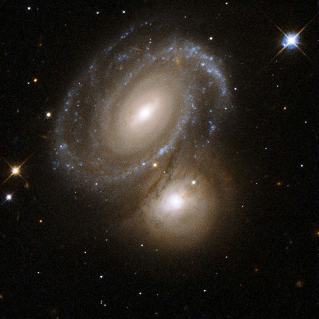 Spiral galaxy above and slightly left of center, blue-white arms made of of stars, and streaks of brown gas. To its lower right, just below central, an orange-white glowing sphere. Bright foreground star in the upper-right.