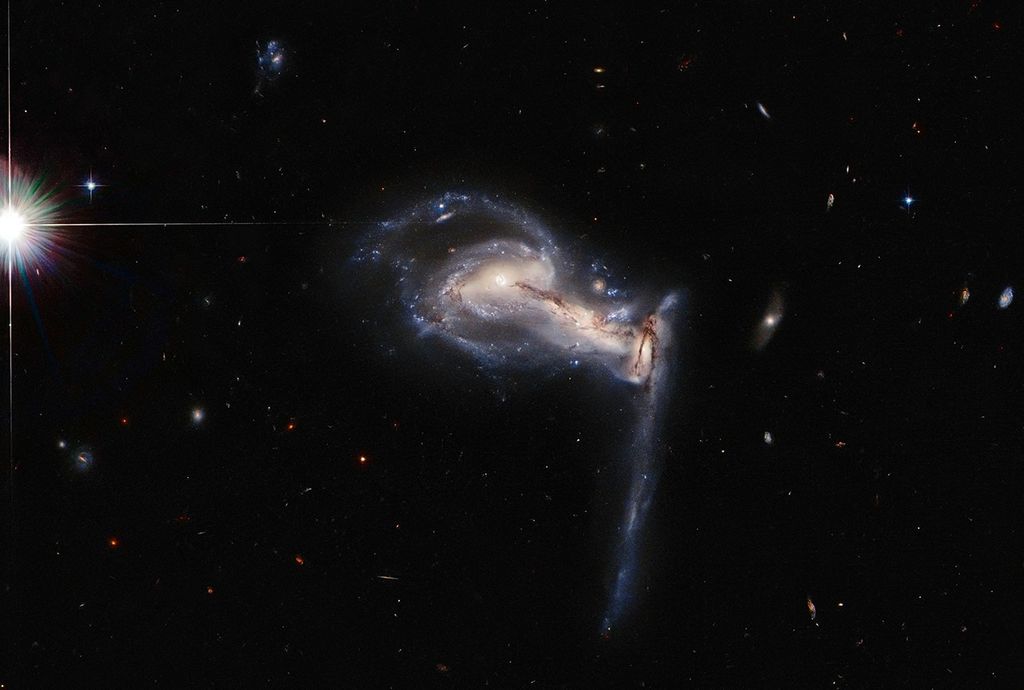Three galaxies, the center one is barely visible and bleeds into the others on the right and left. The one on the right has a long trail of stars stretching toward the bottom. The left one still appears as an intact spiral.. A bright diffracted foreground star at far-left.