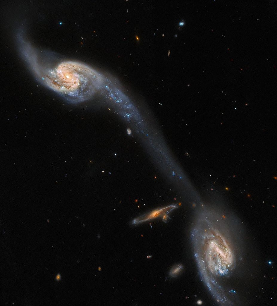 Two face-on spiral galaxies at upper-left and lower-right corners. a long, faint, pale-blue streak joins them, crossing the field diagonally. a small, orange, edge-on spiral galaxy at left of the lower galaxy.