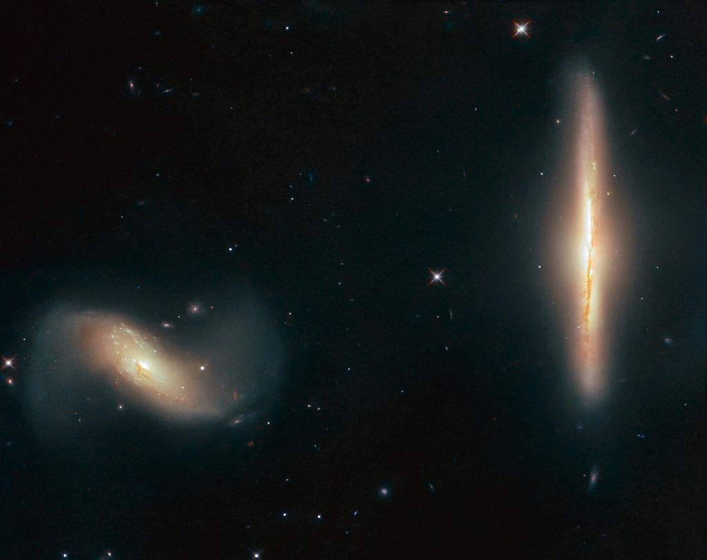 Two galaxies. The one at the right of the image is seen edge-on and vertical, glowing yellow-orange. The one at the left is a tilted disk, white and yellow near the center and reddish and white at the outsides.