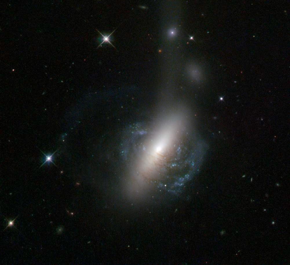Spiral galaxy with blue-white rings of stars and red-brown streaks of gas. The center glows white so brightly that it seems to form a bar above and below the galaxy.