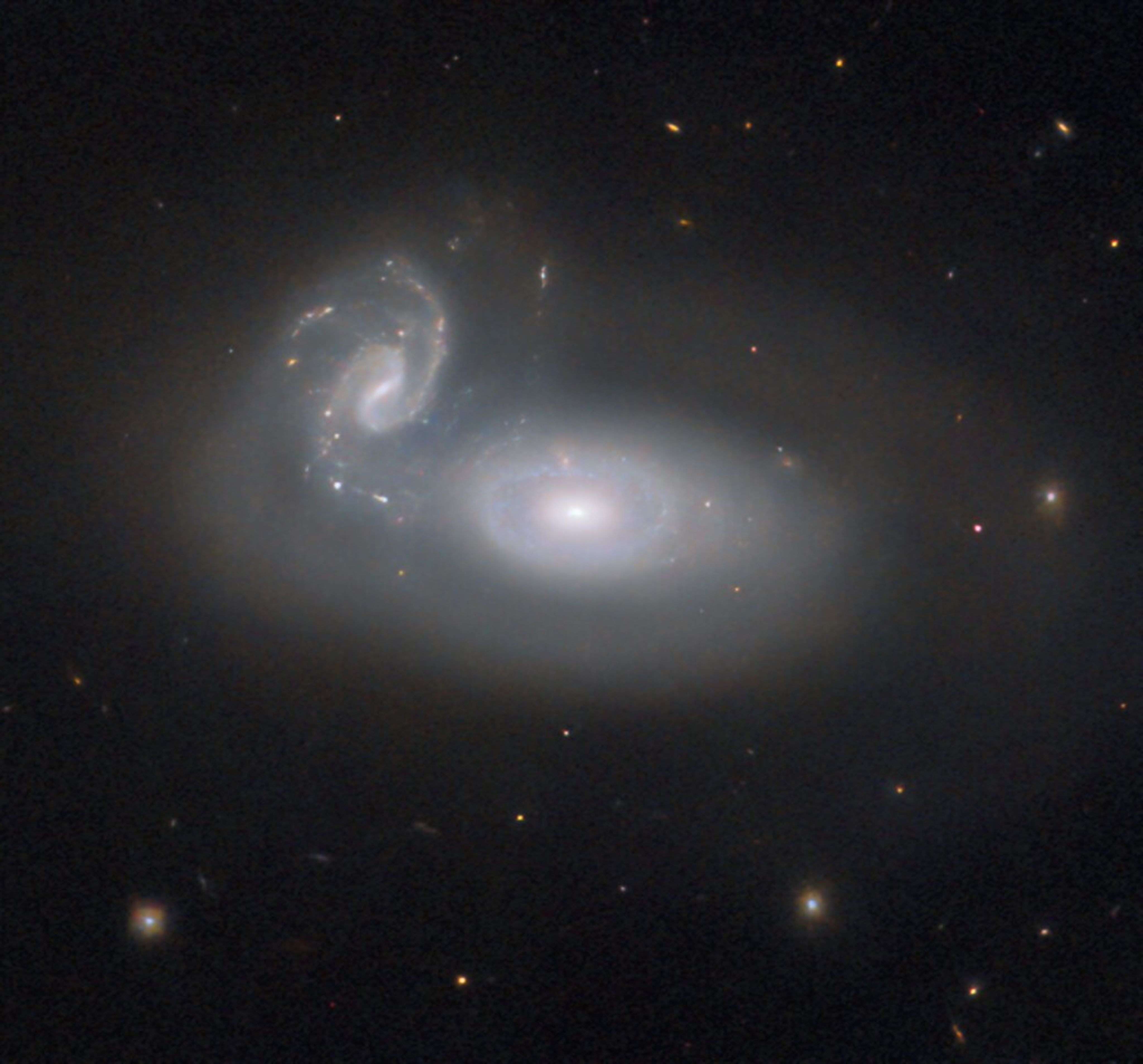 In the center is a large, oval-shaped galaxy, with a shining, ringed core. Left of its center is a second, smaller galaxy with two spiral arms. The galaxy pair is so close that they appear to be merging: a tail of material with a few glowing spots connects from one of the smaller galaxy’s spiral arms to the larger galaxy. A faint halo surrounds both galaxies. Several stars are visible around the pair.