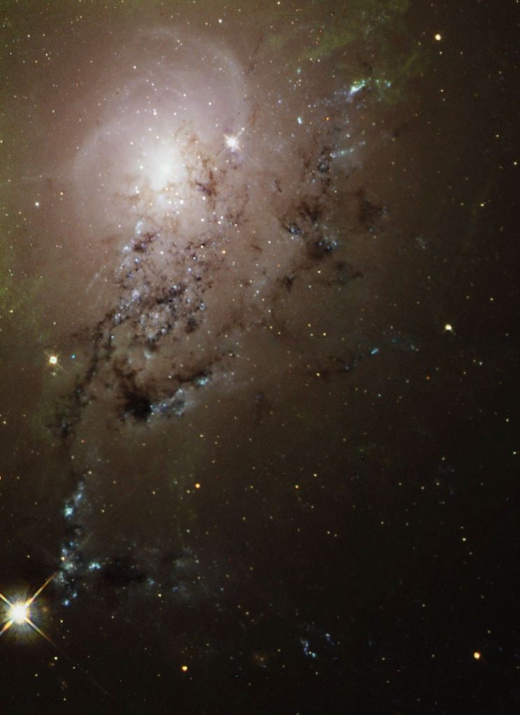 In the upper left is a glowing white core with a pinkish haze around it, and a greenish haze at its edges, particularly the upper left. To its lower right, in the foreground, are trails made by "arms" of white stars and black gas/dust.