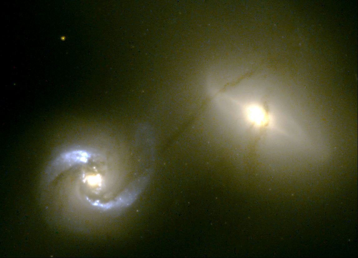 Two bright galaxies with glowing yellowish cores. The one at bottom-left has two thick blue-white spiraling arms, and the one at center right has a thin glowing white haze around it. Both of them seem to be surrounded by a yellow glow.,