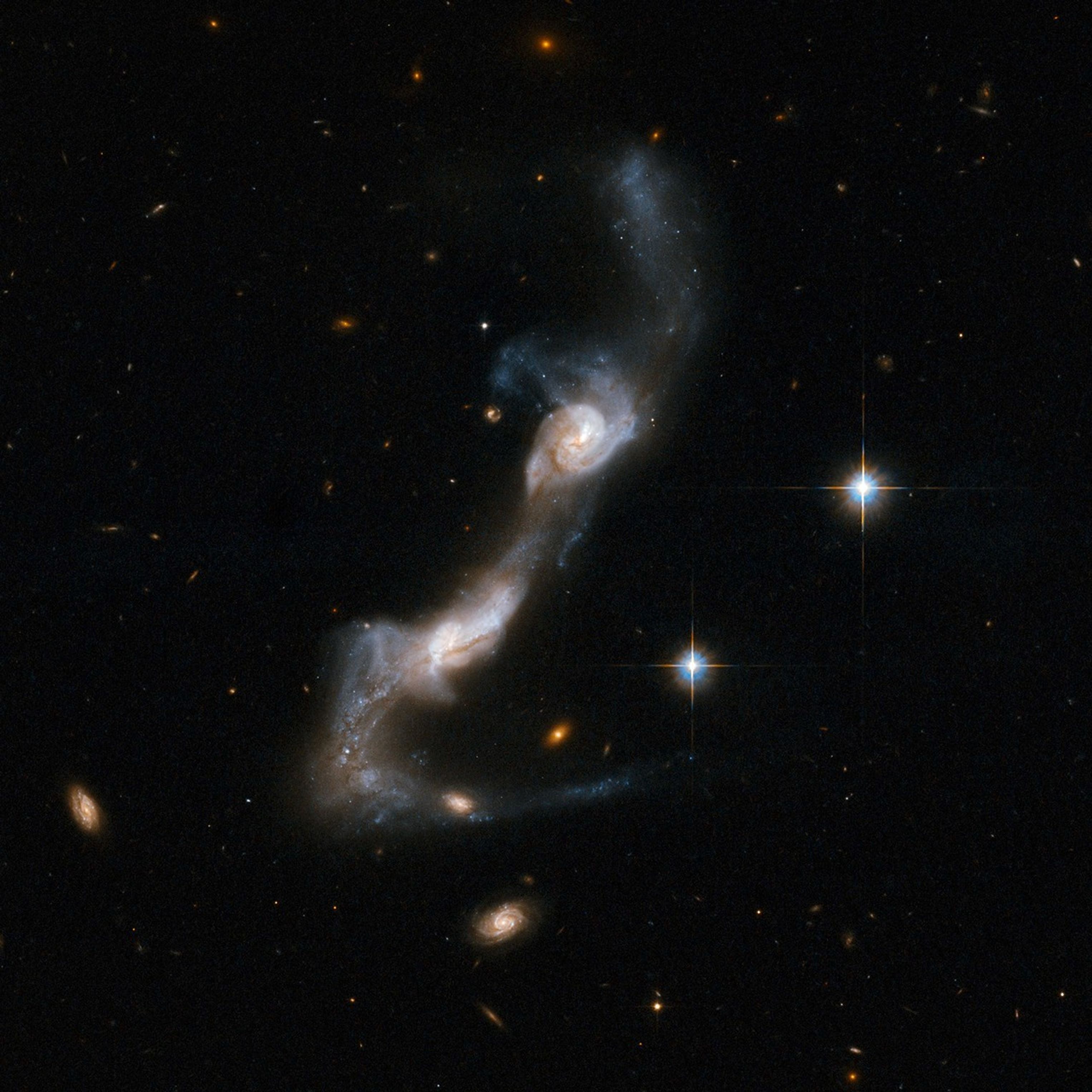 Two distorted galaxies, looking like white-and-orange shells with streaks of white stars. The galaxies are above-right and below-left compared to each other. More spiral galaxies in the background near the bottom, two diffracted foreground stars on the right.