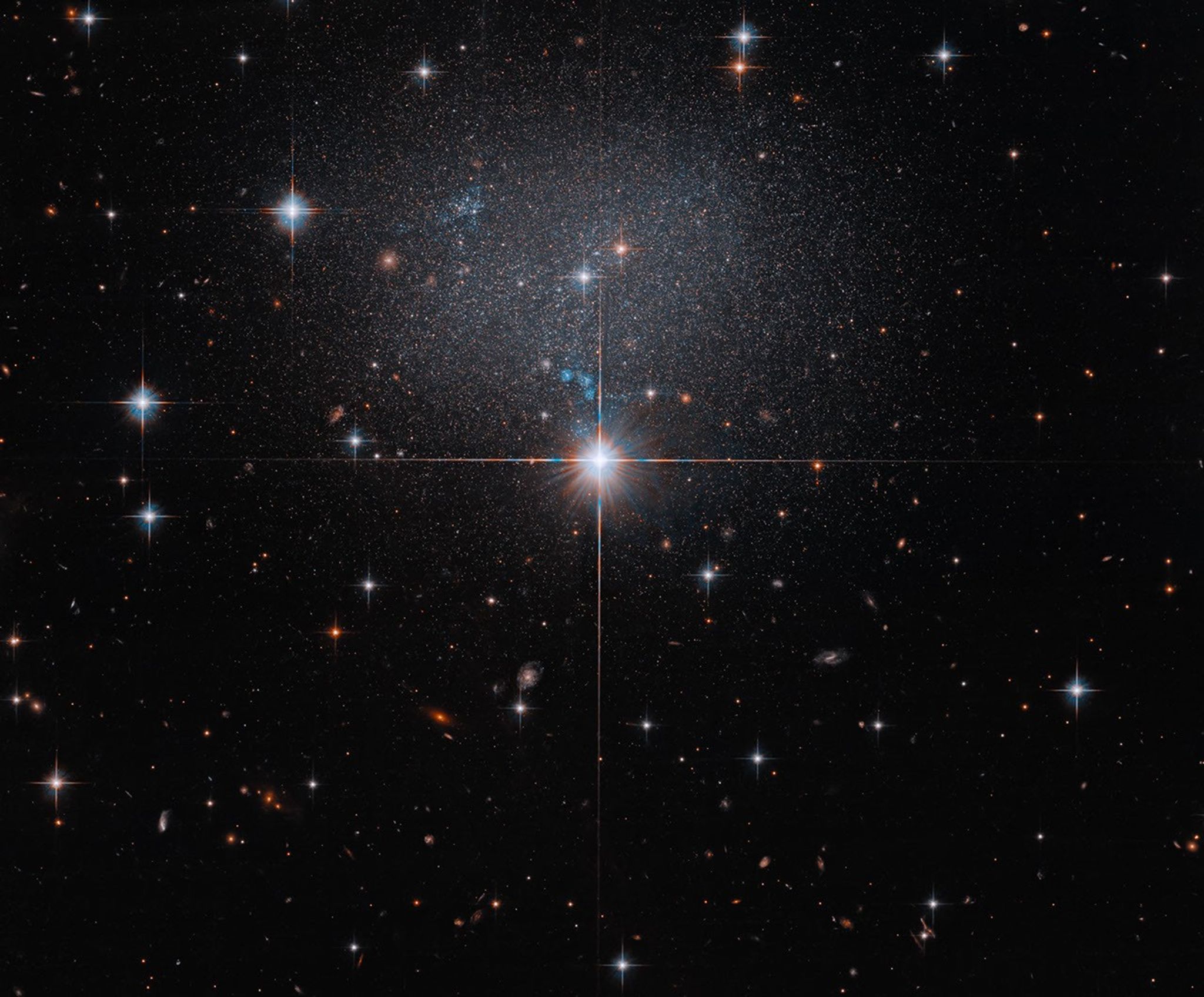 A foreground star at image center shines brightly with four diffraction spikes. Above this star is a diffuse and faint dwarf irregular galaxy. The galaxy itself has bright blue and red stars.