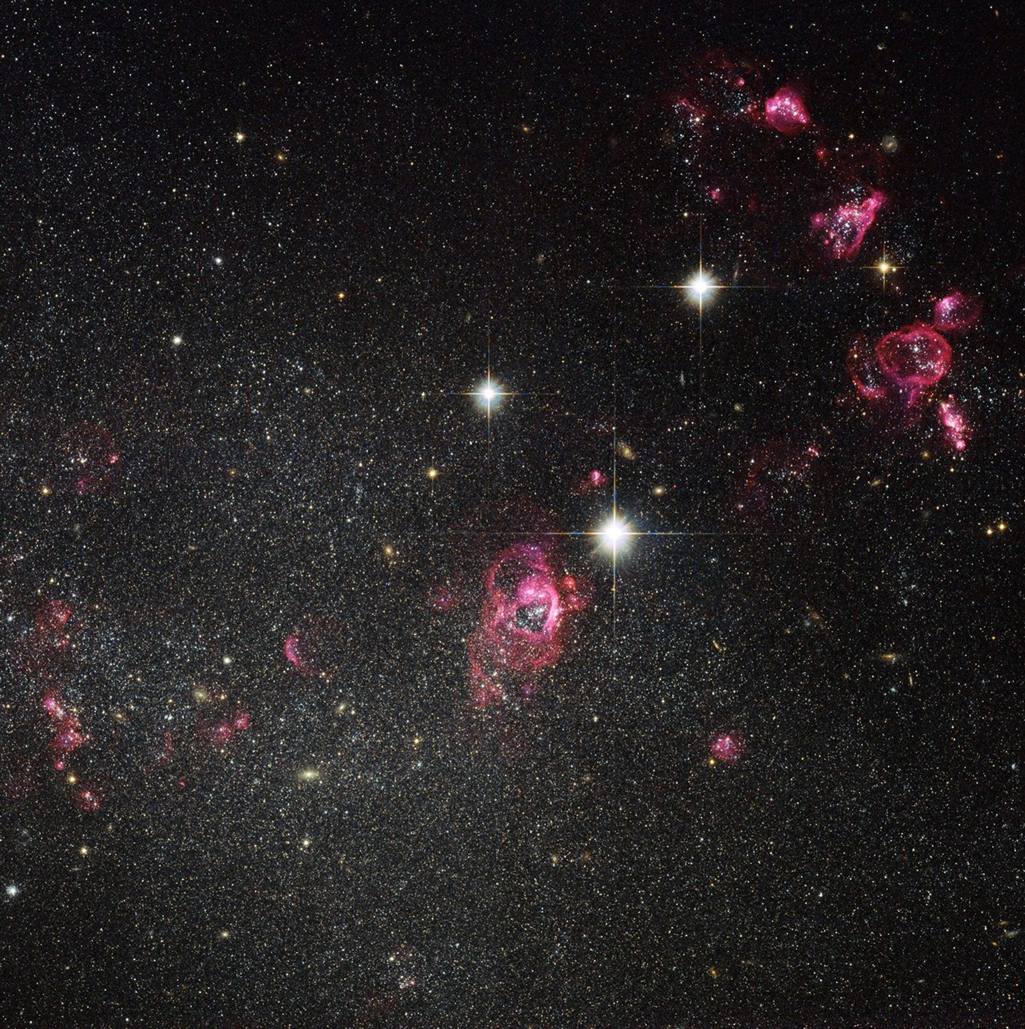 Near the center of the image, a reddish-pink ring-shaped cloud of gas. Above-right are three white foreground stars, and in the top-right corner, three more reddish-pink ring-clouds. Thousands of white stars and a few more red-pink clouds dot the background.