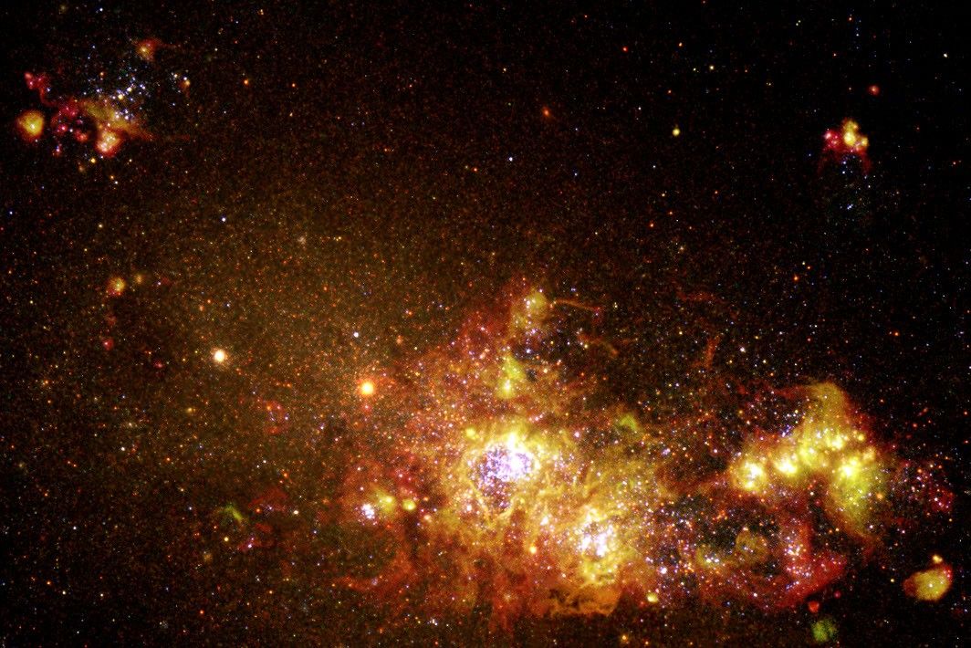 The nebula appears in the foreground in the lower right as a glob of red, orange, and yellow gas and dust with glowing white stars within, and smaller globs of red and yellow gas and dust in the upper left and upper right. The dwarf galaxy appears as a fuzzy collection of yellow-orange stars in the background on the left.