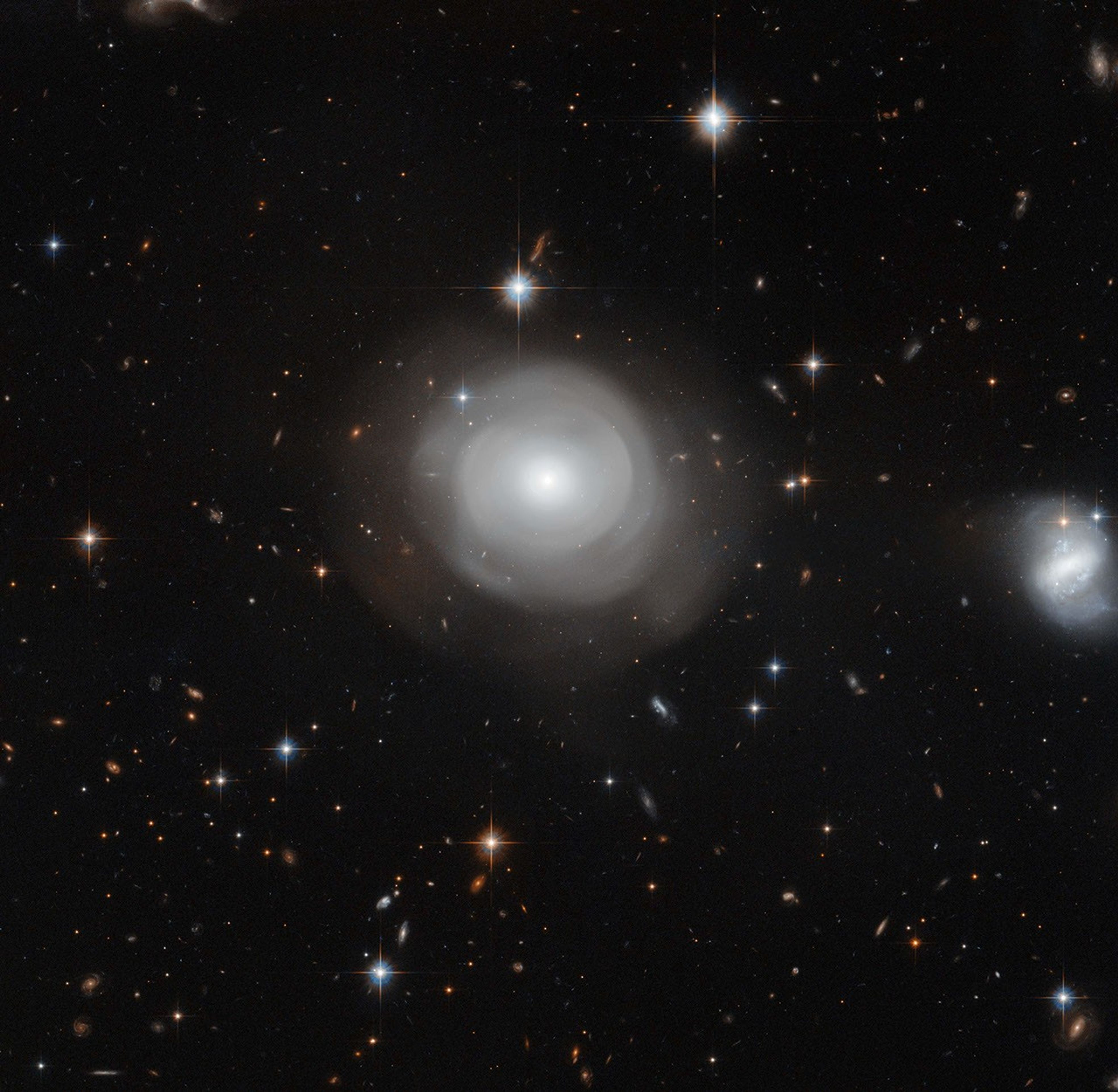 Bright, white diffuse sphere with an extremely bright core. Diffuse shells of matter form layers around the core. A small blue-white spiral lies at the right edge of the image. Hundreds more galaxies of different shapes and colors shine in the background.