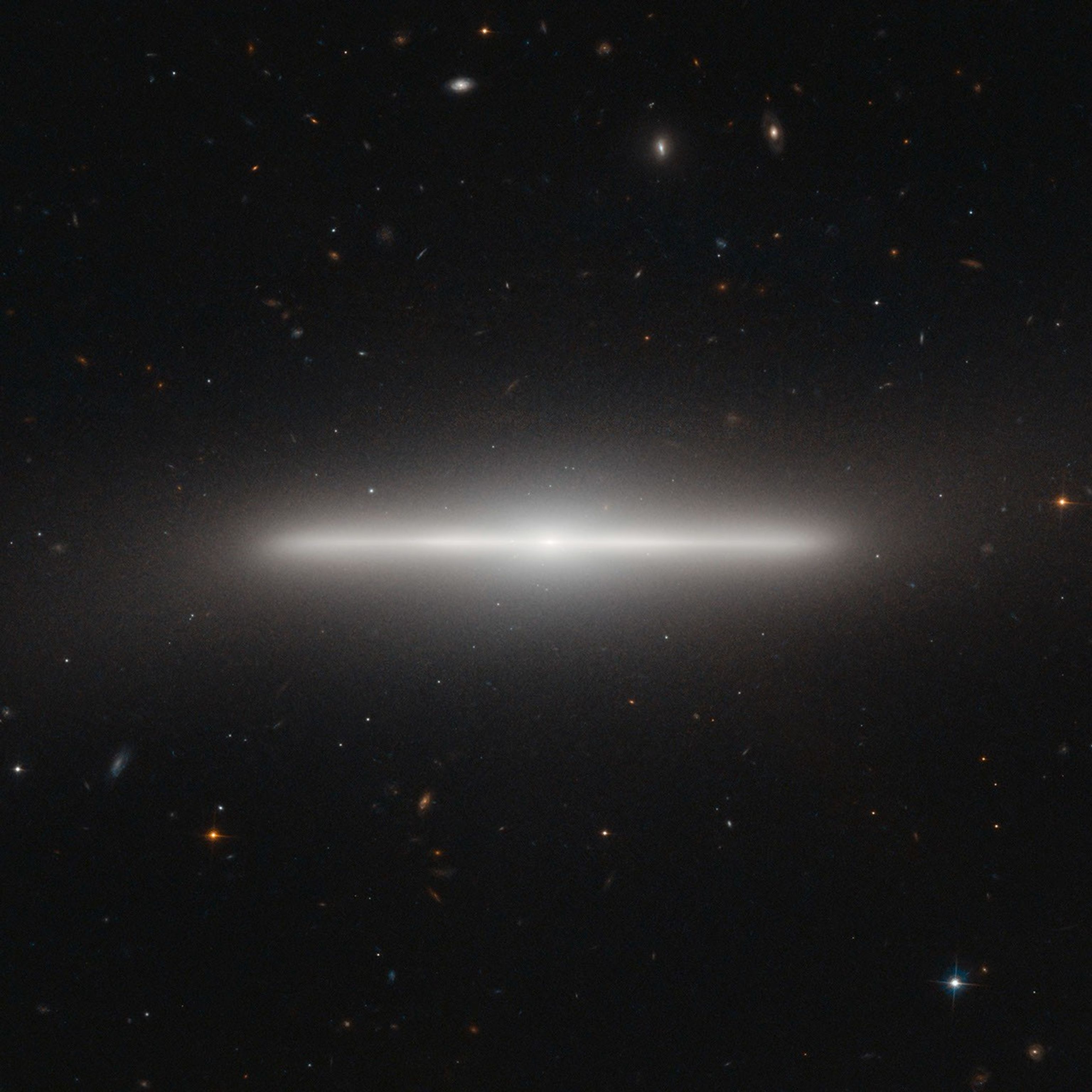 The edge-on galaxy looks like a white "line" in space with a glowing white halo around it. Hundreds more galaxies and stars of different shapes and colors dot the black background.