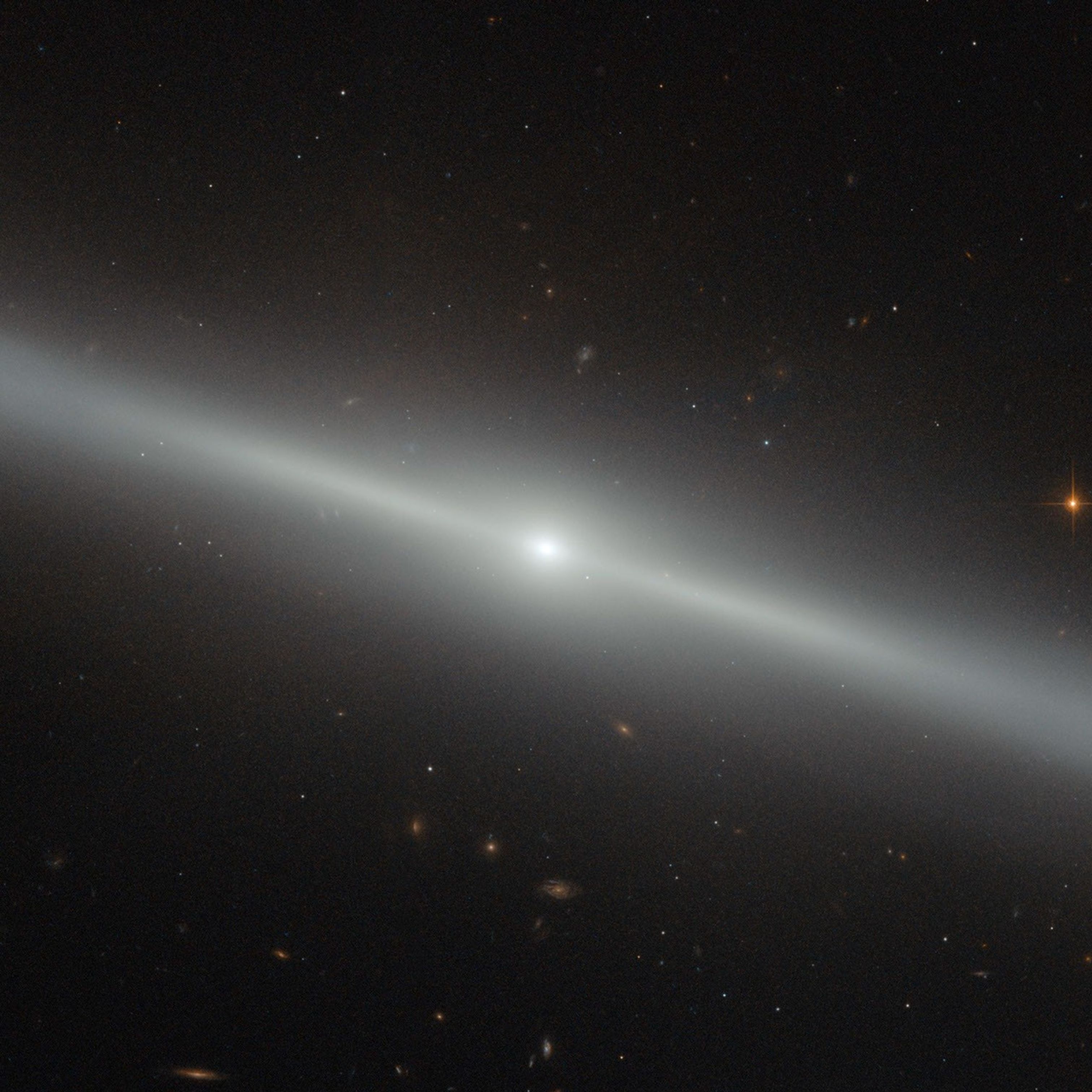 The edge-on galaxy looks like a white "line" in space with a glowing white halo around it, stretching across the entire image at a slight angle. Dozens more galaxies and stars of different shapes and colors dot the black background.