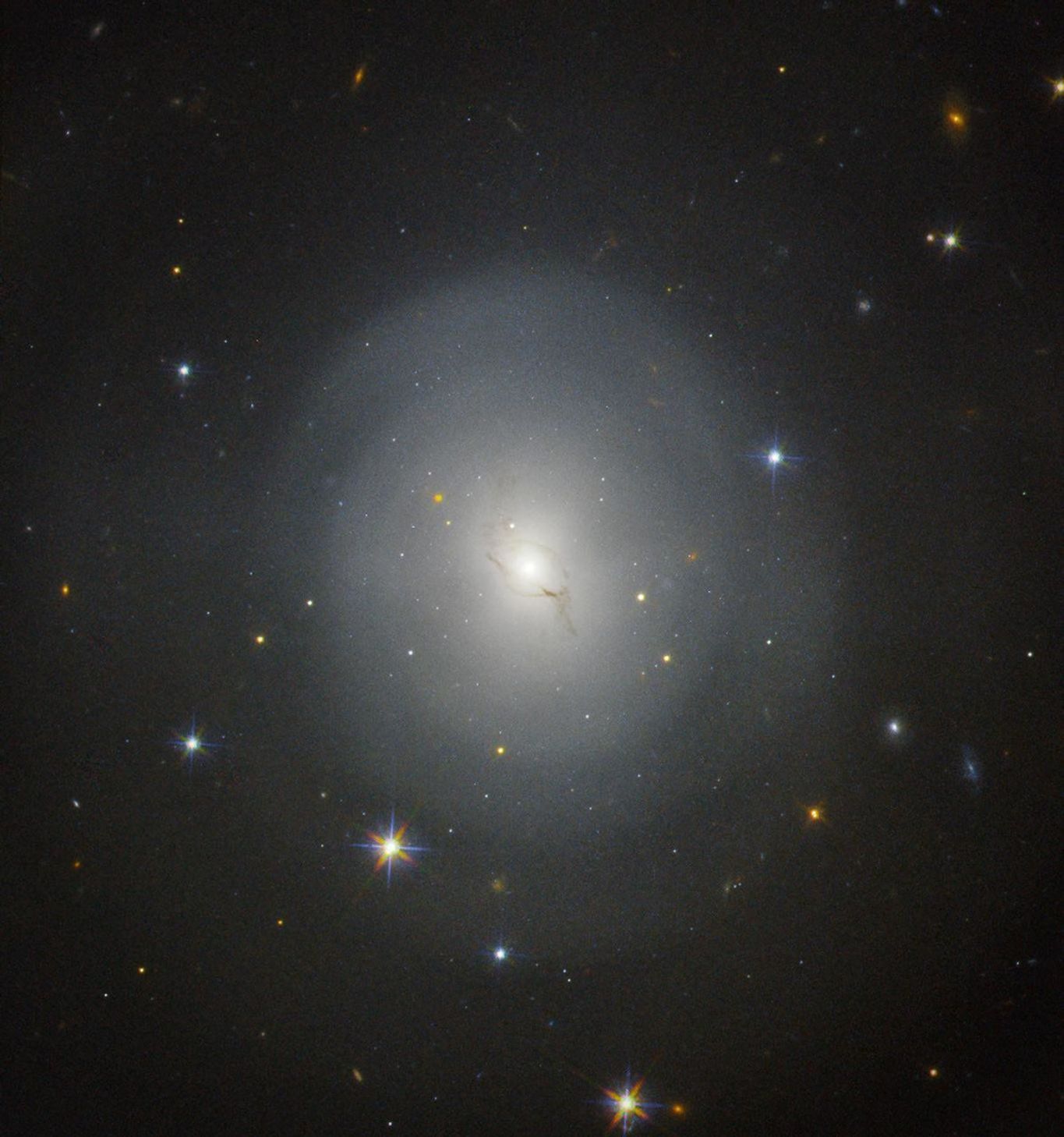 A galaxy in the center of the image glows bright white and is circled by a few trails of dark dust and gas. Many more stars and galaxies shine brightly in the background against the black backdrop of space.