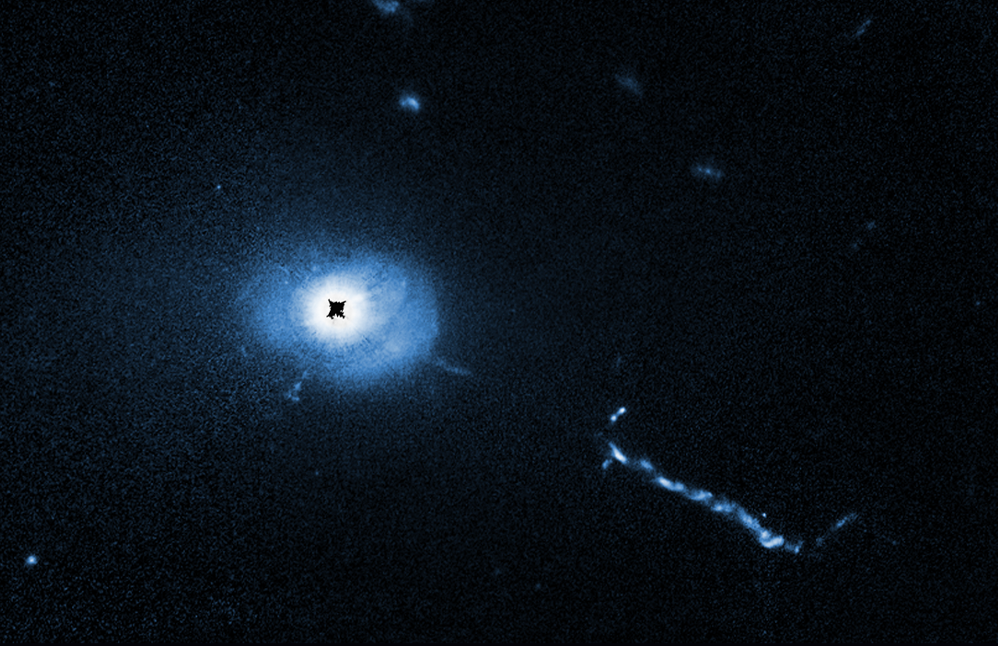 A Hubble Space Telescope Imaging Spectrograph (STIS) coronagraph image of quasar 3C 273. It looks the same as the WFPC2 image, but in shades of blue. A black circle (labeled “core light blocked”) blocks the glare of the quasar. Blue-colored filamentary material is visible near the black hole. The extragalactic jet is still visible.