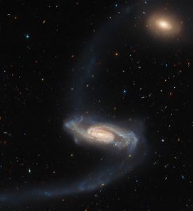 A spiral galaxy. It has a bright core with patches of dark dust, and fuzzier, dimmer spiral arms in cooler colors, with spots of bright blue. Long, faint tidal streams stretch from the galaxy’s arms: one up to the top of the frame, one curving down to the bottom-left corner. In the top-right there is a smaller, orange elliptical galaxy. The background is studded with many tiny stars and galaxies.
