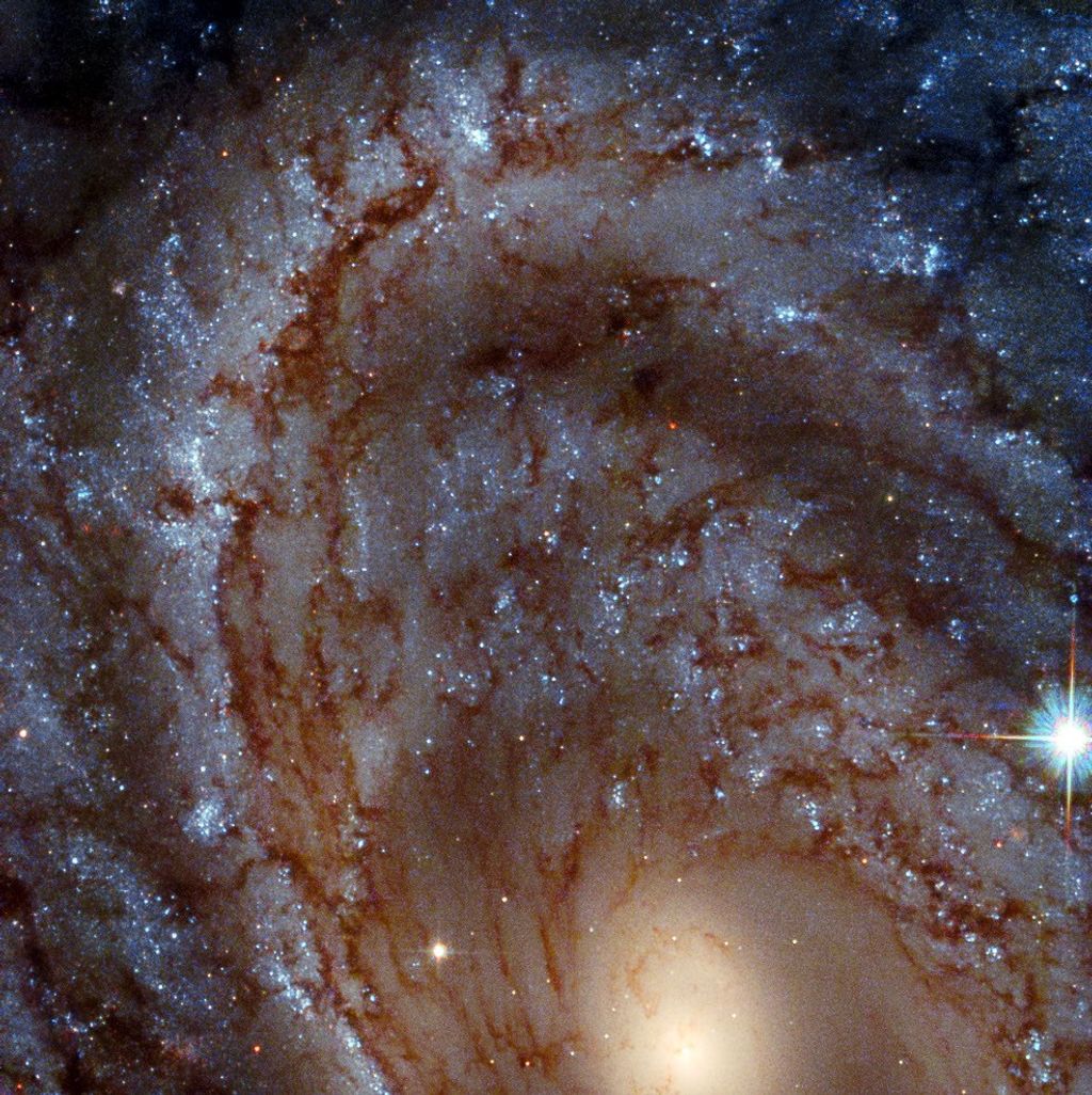 A spiral galaxy’s whirling arms fill the image. The yellow galactic core is visible at the bottom center of the image, with arms arcing upward. Dark-brown dust and bright-blue stars lace through the spiral’s arms. A foreground star with diffraction spikes shines at the lower-right side of the image.