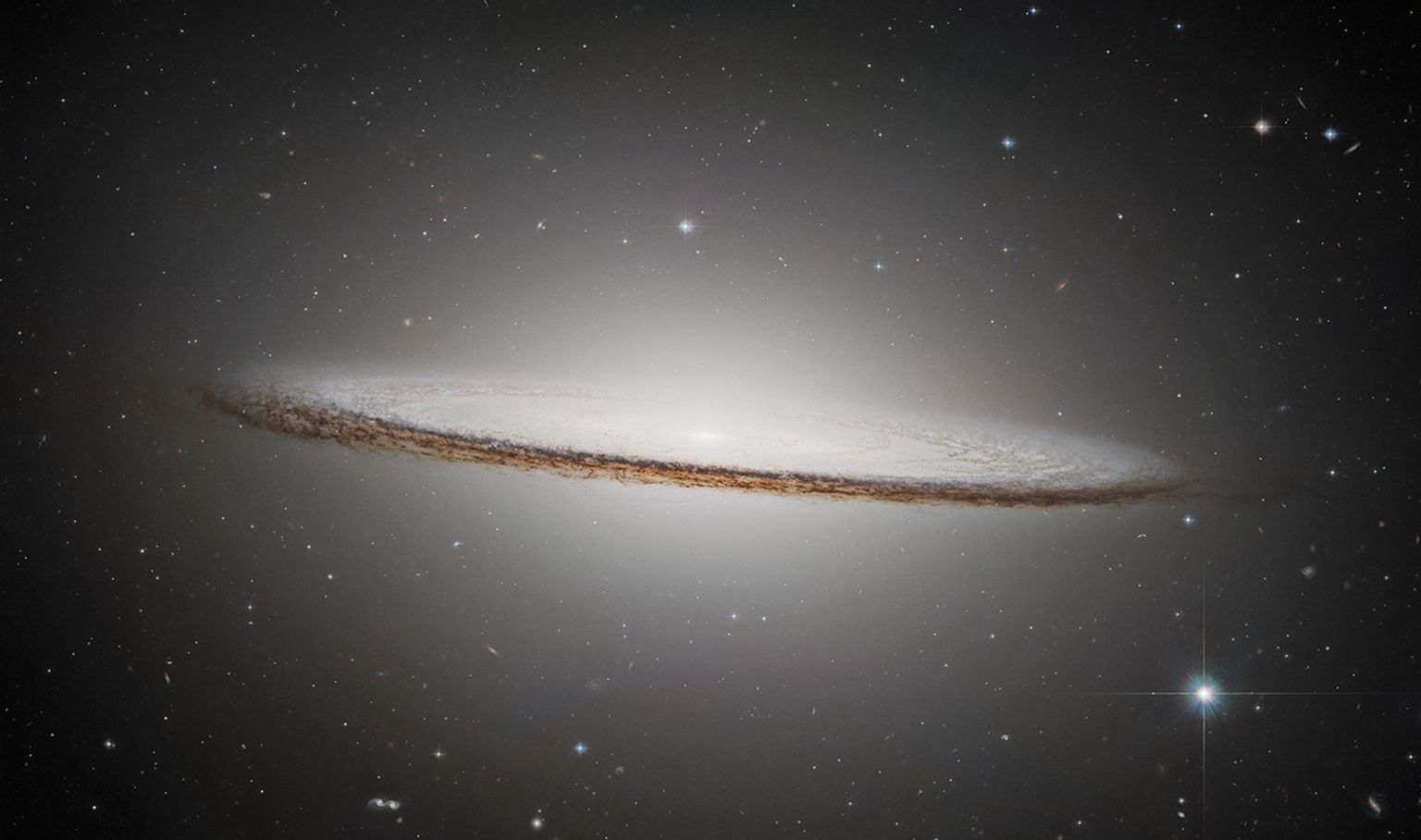 The Sombrero Galaxy is an oblong, pale white disk with a glowing core. It appears nearly edge-on but slants slightly in the front, presenting a slight top-down view of the inner region of the galaxy and its bright core. The outer disk is darker with shades of brown and black. Different colored distant galaxies and various stars are speckled among the black background of space surrounding the galaxy.