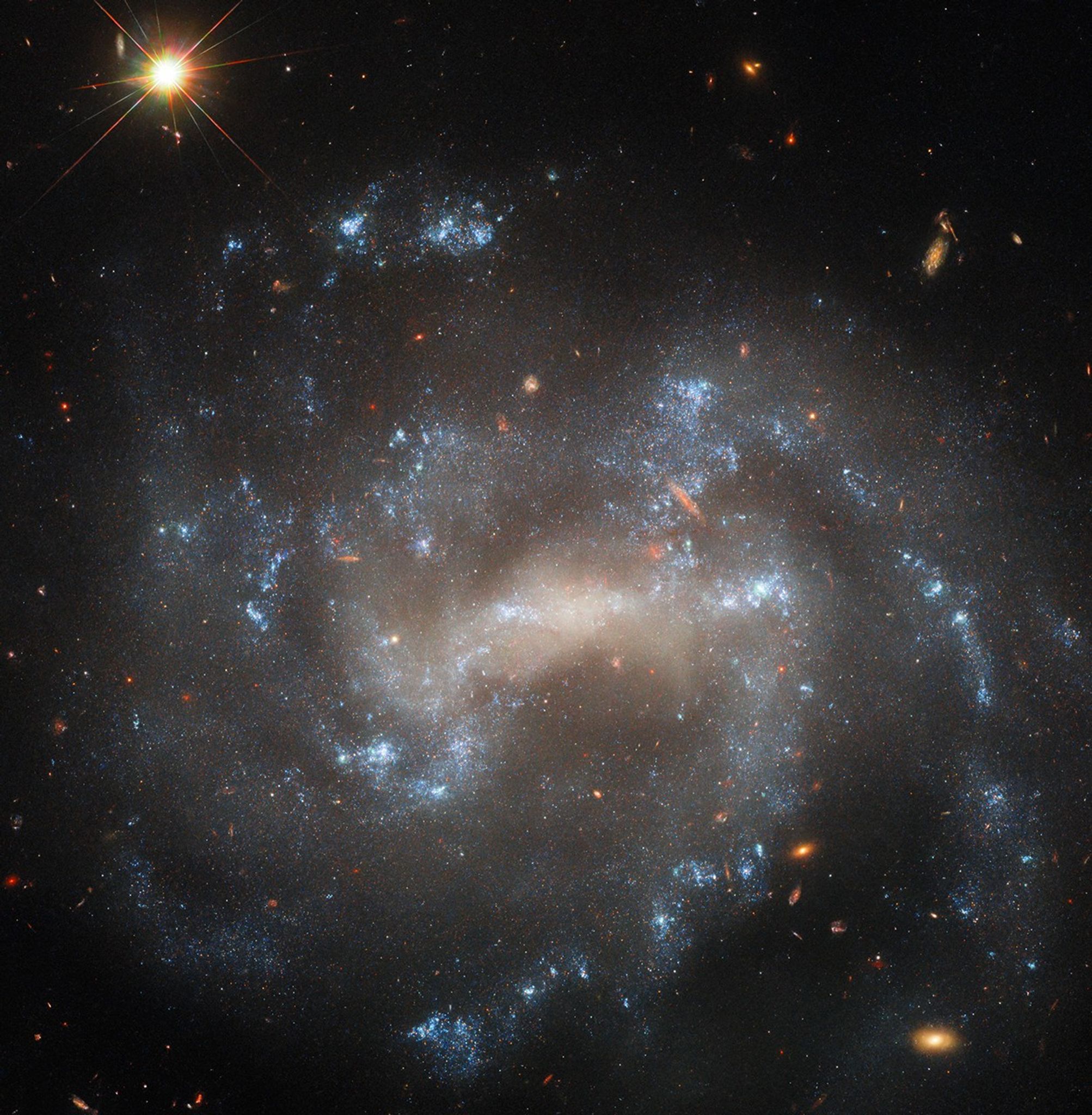 A spiral galaxy seen close to face-on. The center of its disk is a bright, pale yellowish, oval shape. Spiral arms extend from either side of the oval through the disk on irregular paths. The arms hold bright bluish-white patches of stars that mark their extent. Distant background galaxies appear as small orangish blobs around the spiral galaxy. In the top-left corner, a nearby star shines brightly, diffraction spikes are radiating from it.