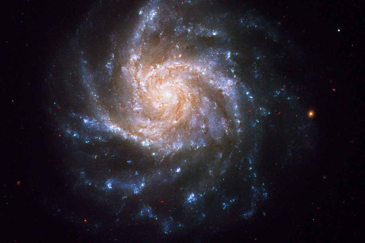 A spiral galaxy with a glowing yellow core, pinwheel arms of stars in pink and blue-white, and thinner swirls of orange-brown dust. The galaxy is set against the black background of space, with dozens of distant galaxies and stars.