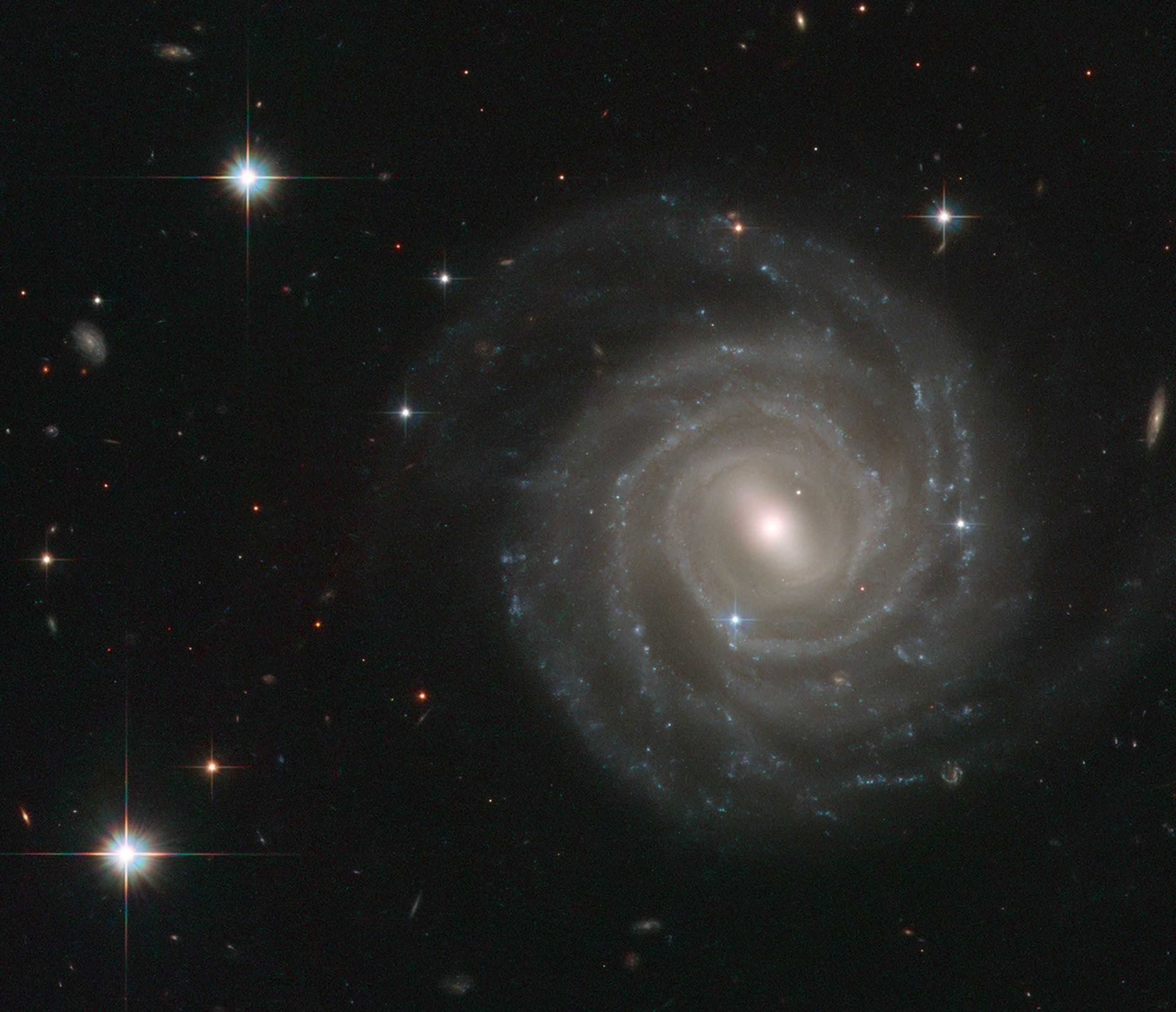 The majestic galaxy right-of-center has a pinwheel shape made up of bright blue stars wound around a yellow-white hub of central stars. The hub has a slash of stars across it, called a bar. The galaxy is tilted face-on to our view from Earth. A slightly s-shaped white line across the bottom-right is an asteroid streaking across Hubble's view. It looks dashed because the image is a combination of several exposures of the asteroid flying by like a race car. Three foreground stars shine in the upper-left, lower-left, and above-right of the galaxy.