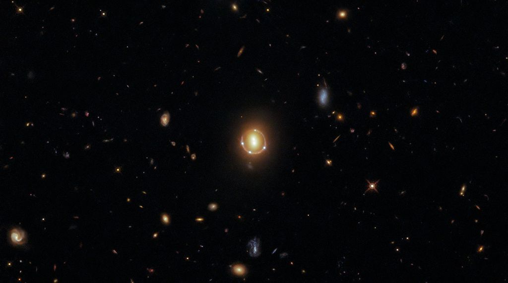 Black background dotted with galaxies. Two reddish galaxies are next to each other as two vertical dots at image center with a reddish-orange ring curving almost completely around it, with four white dots around the ring.