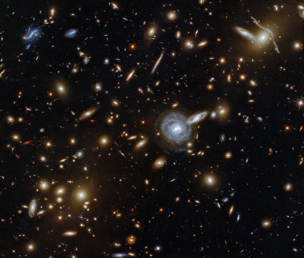 A cluster of various types of galaxies, visible as a dense crowd of oval shapes. Two spiral galaxies are especially prominent near the center, and another in the upper right, where the lensing effect is best visible.