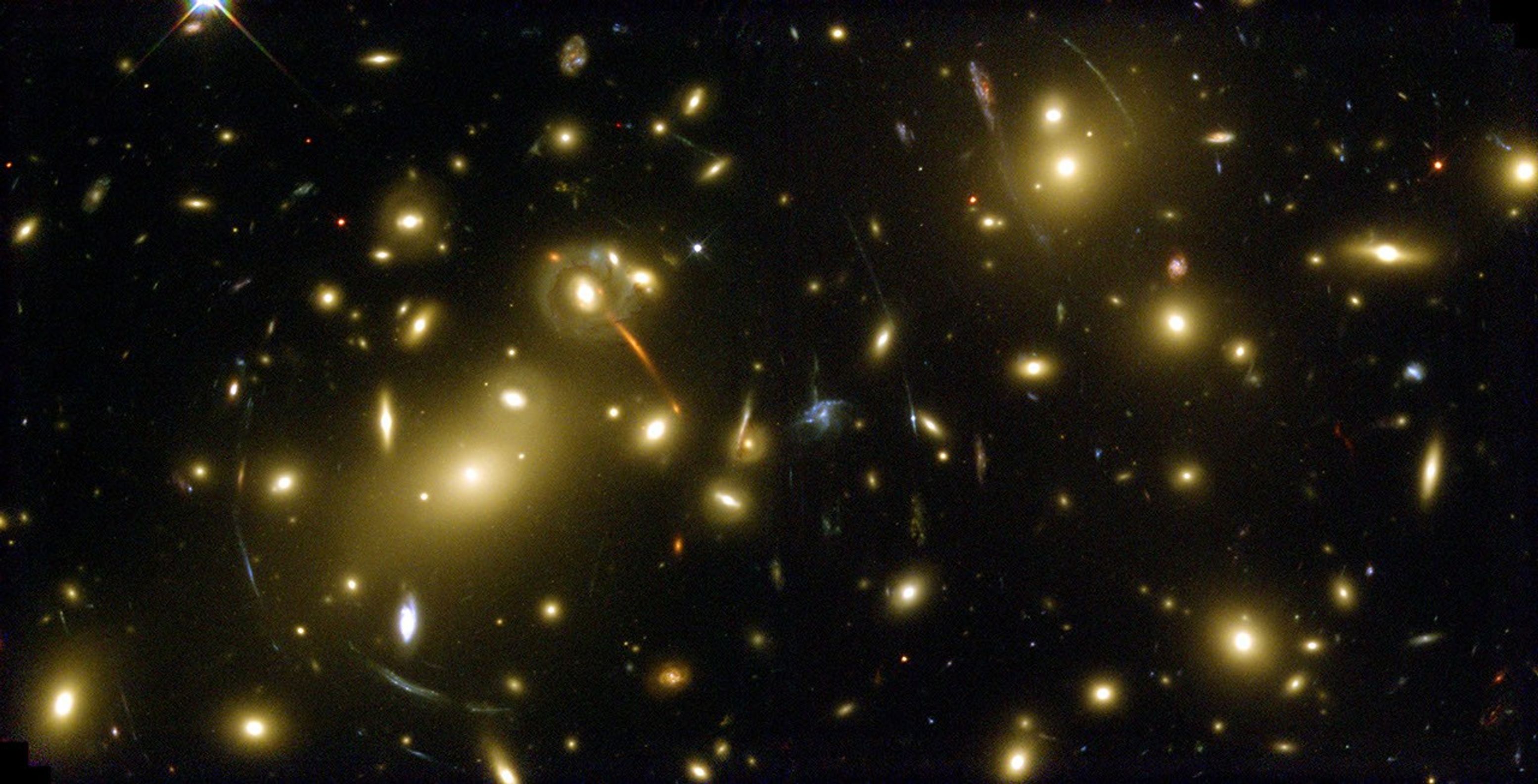 Bright yellow-white galaxies fill the field of view. Faint streaks and arcs of gravitationally lensed background galaxies.