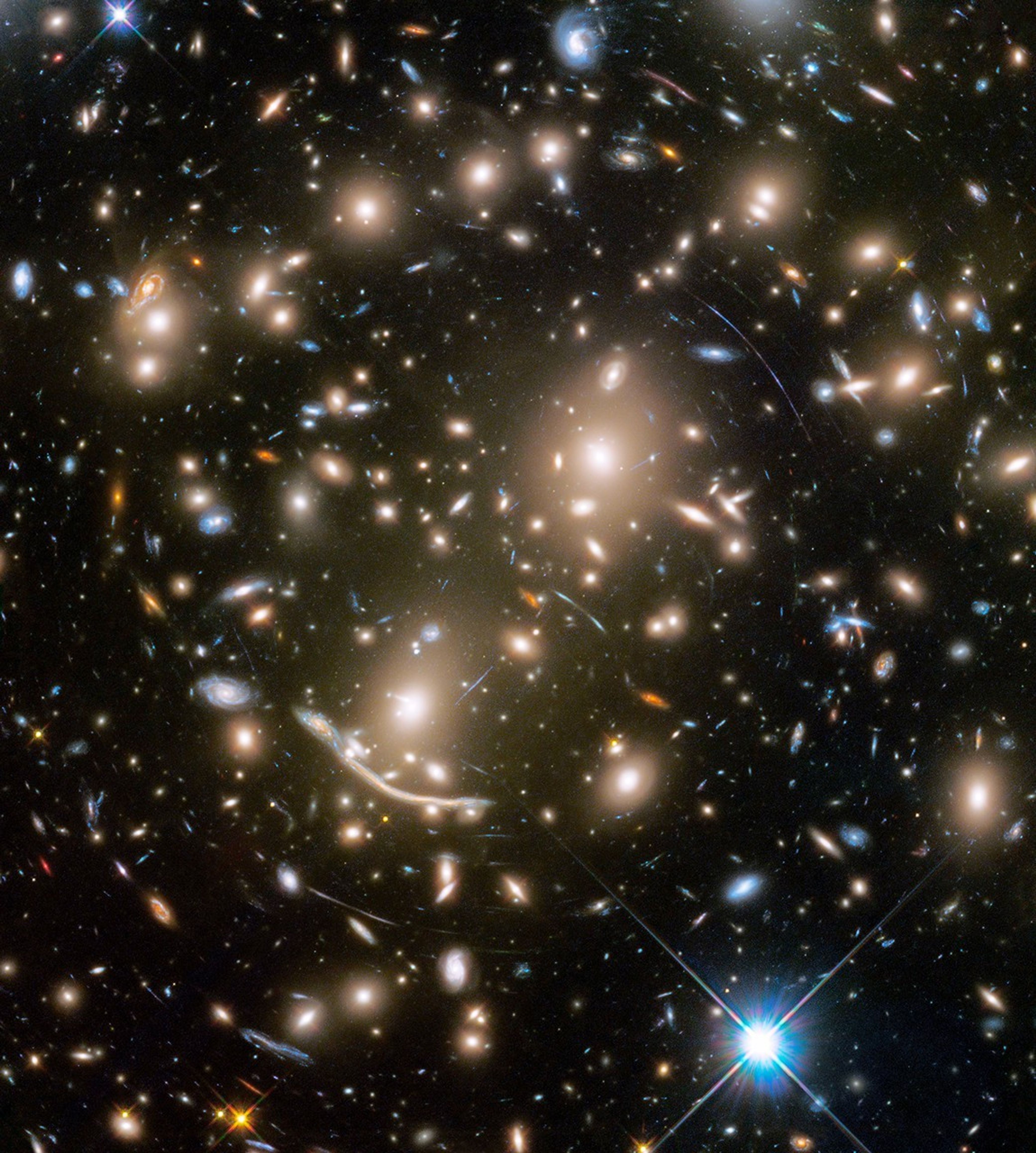 A packed field of galaxies and curved blue streaks and arcs. Large grouping of galaxies warped around what appears to be a giant lens. Each galaxy appears as a glob of light. Bright blue-white foreground star at bottom right.