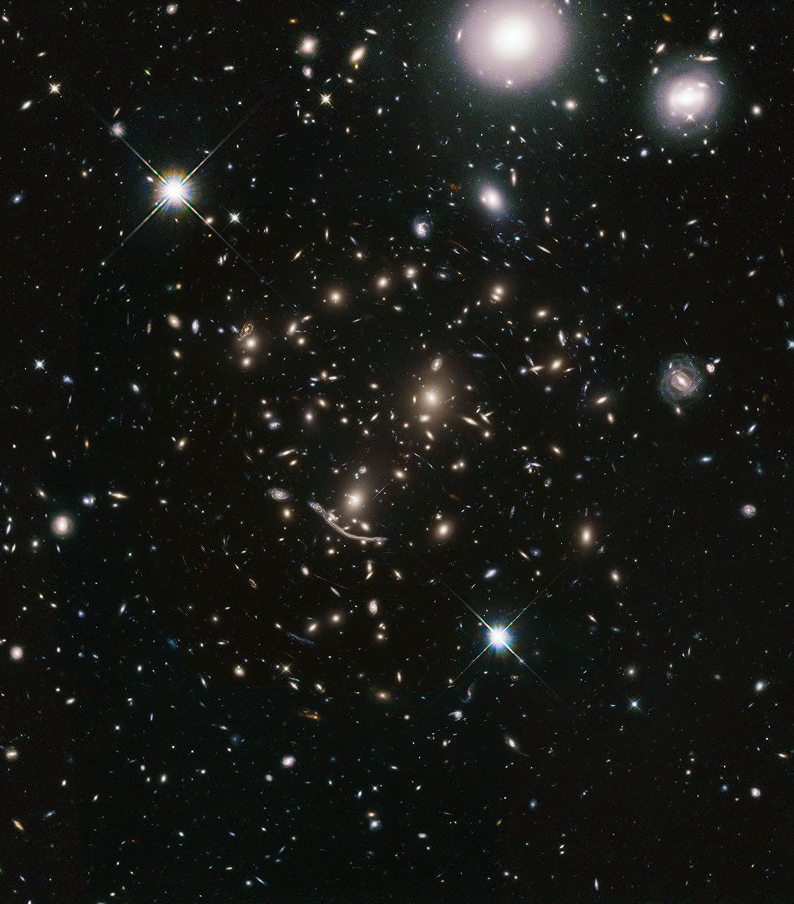 A packed field of galaxies and curved blue streaks and arcs. Large grouping of galaxies warped around what appears to be a giant lens. Each galaxy appears as a glob of light. Bright blue-white foreground star at lower-center and upper left, and bright white foreground galaxies at upper-right.