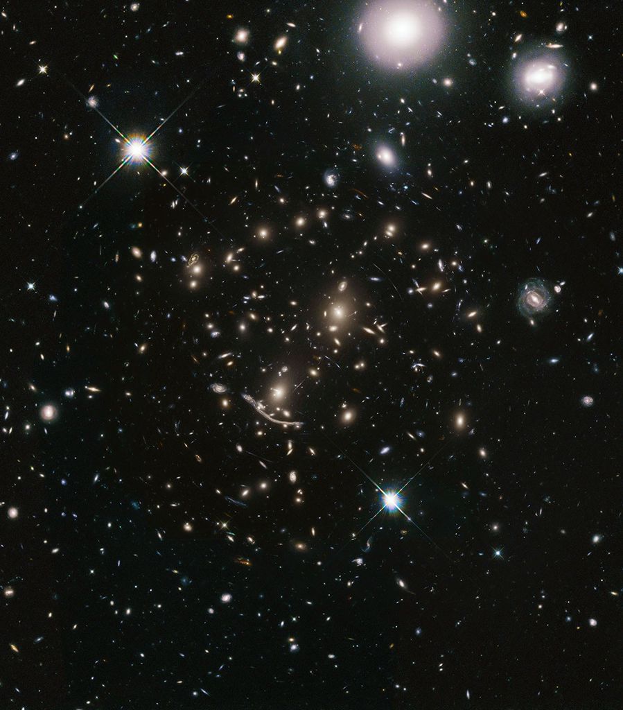 A packed field of galaxies and curved blue streaks and arcs. Large grouping of galaxies warped around what appears to be a giant lens. Each galaxy appears as a glob of light. Bright blue-white foreground star at lower-center and upper left, and bright white foreground galaxies at upper-right.