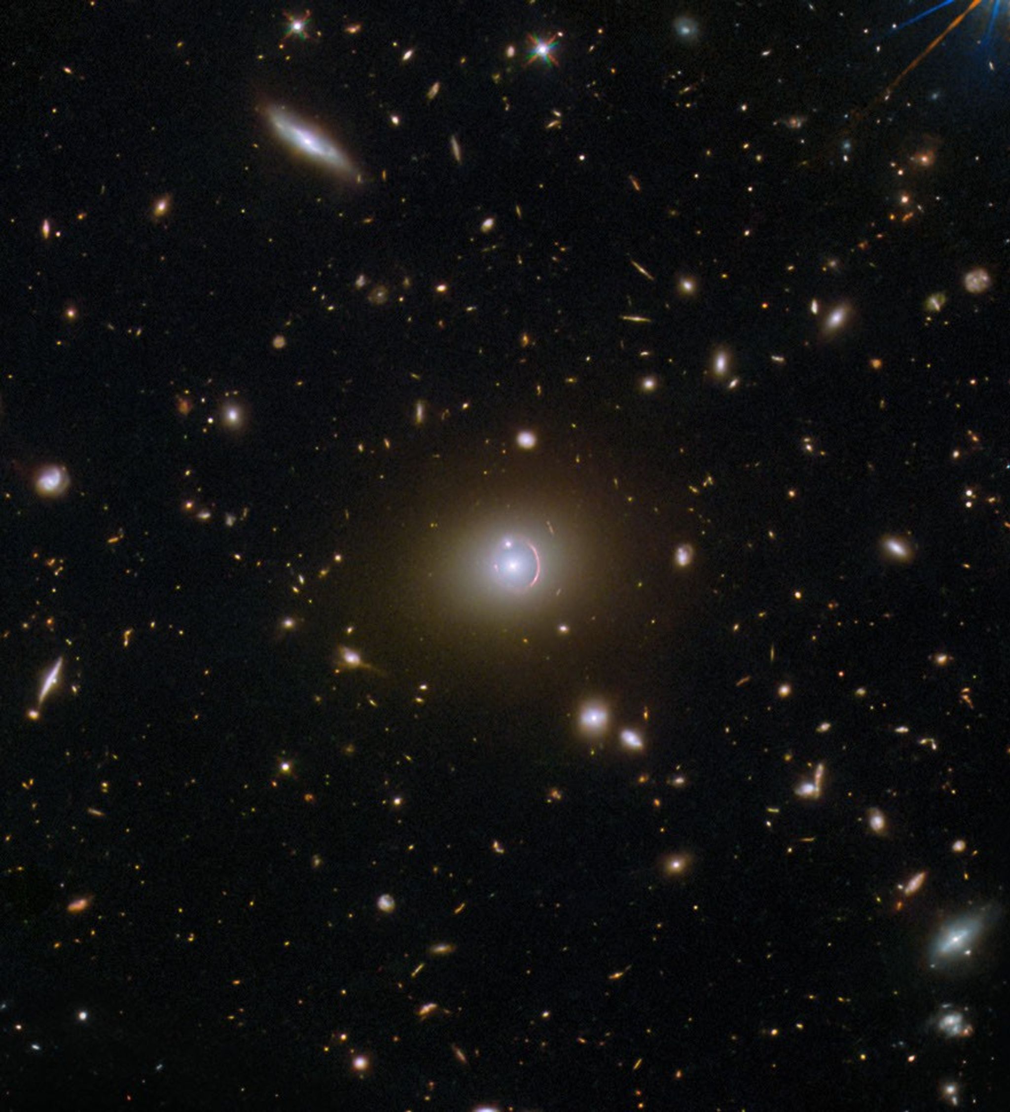 A field full of distant galaxies on a dark background. Most of the galaxies are very small, but there are a few larger galaxies and some stars where detail is visible. In the very center is an elliptical galaxy with a brightly glowing core and a broad disk. A reddish, warped, ring of light, thicker at one side, surrounds its core. A small galaxy intersects the ring as a bright dot.
