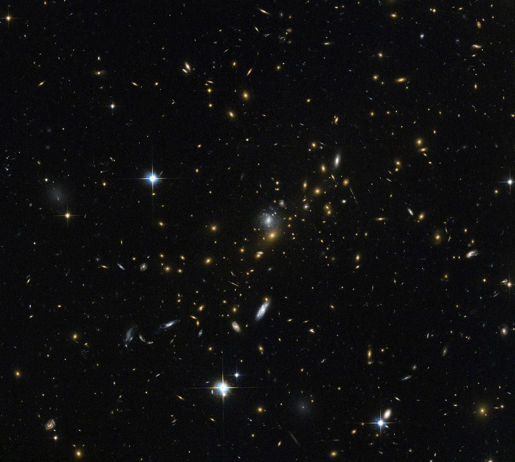 A field of galaxies and with faint curved arcs. Each galaxy appears as a glob of light, most of them reddish-orange. Bright, blue-white, twinkling foreground stars are scattered in a few places in the image.