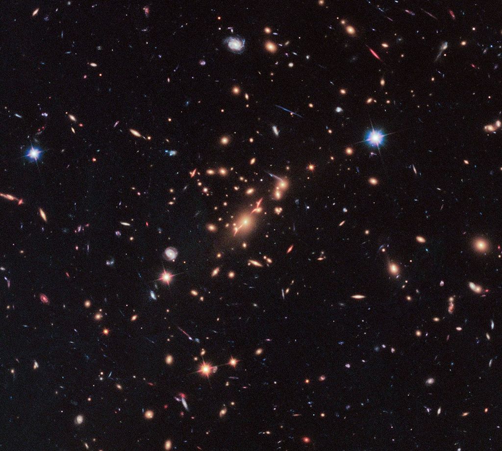 A bunch of bright dots, spheres, and spirals of light (mostly red-orange, with some blue and white) against the black backdrop of space, with a clump near the center that have the circular lensing effect. A bright blue foreground star in the upper-right.