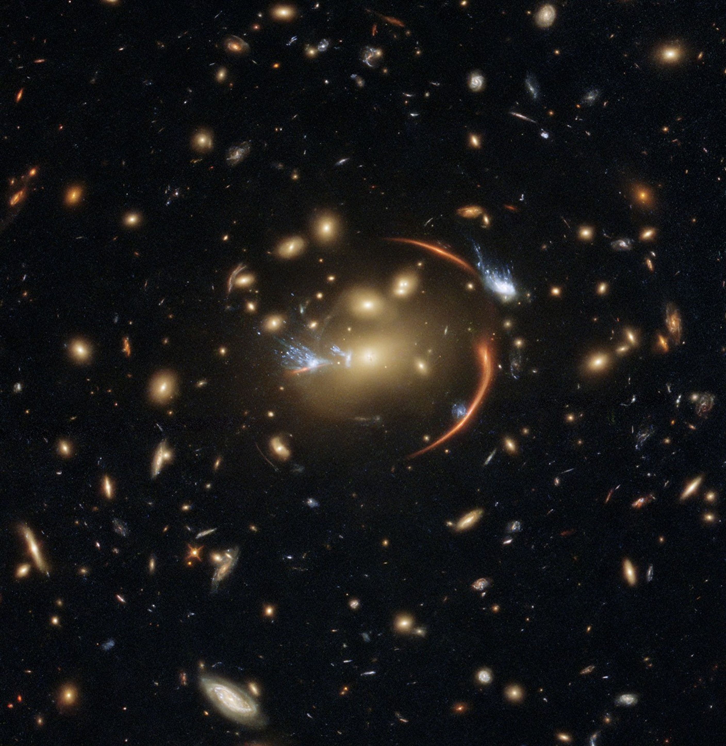 A field full of distant galaxies on a dark background. Most of the galaxies appear small, but there are a few larger galaxies and some stars where detail is visible. In the very center is an elliptical galaxy with a brightly glowing core and a broad disk. A reddish, warped, ring of light, mainly made up of two arcs on the right side, surrounds its core, as do two wispy-white galaxies (one on the left, one on the upper right) that seem to have bluish "tails" flowing out behind them.