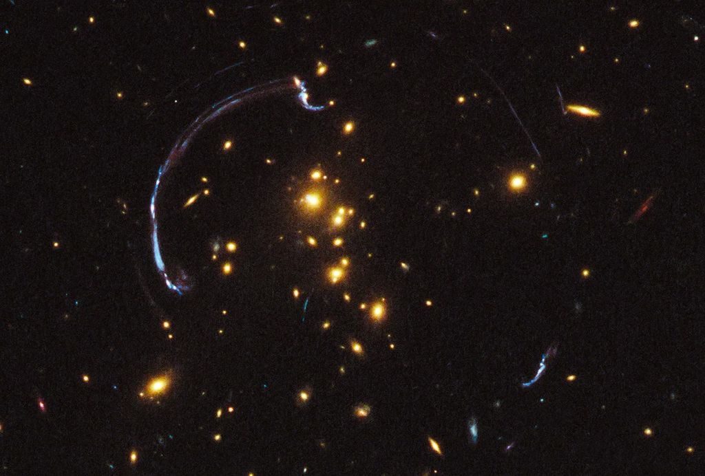 Bright yellow-white galaxies against the black backdrop of space.. Faint streaks and arcs of gravitationally lensed background galaxies.
