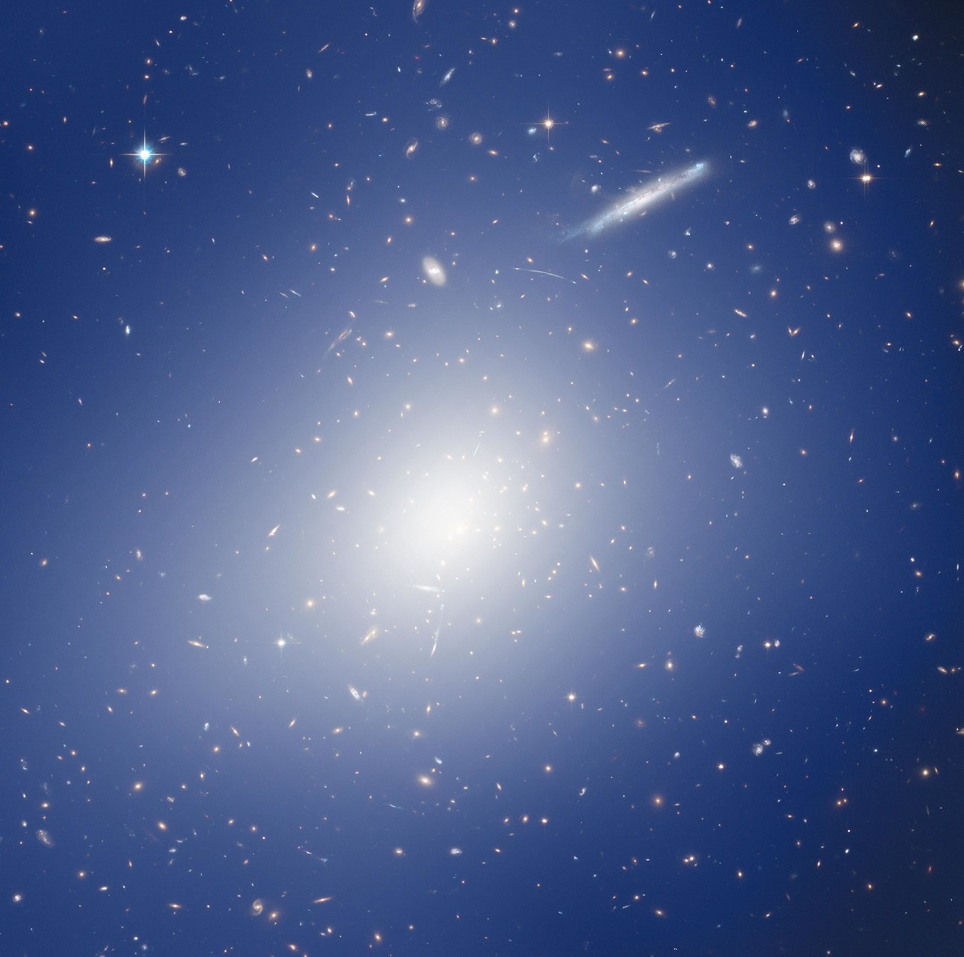 A massive galaxy cluster full of galaxies of all shapes and sizes, which seem to be surrounded by a bluish haze coming from a bright glowing white center.