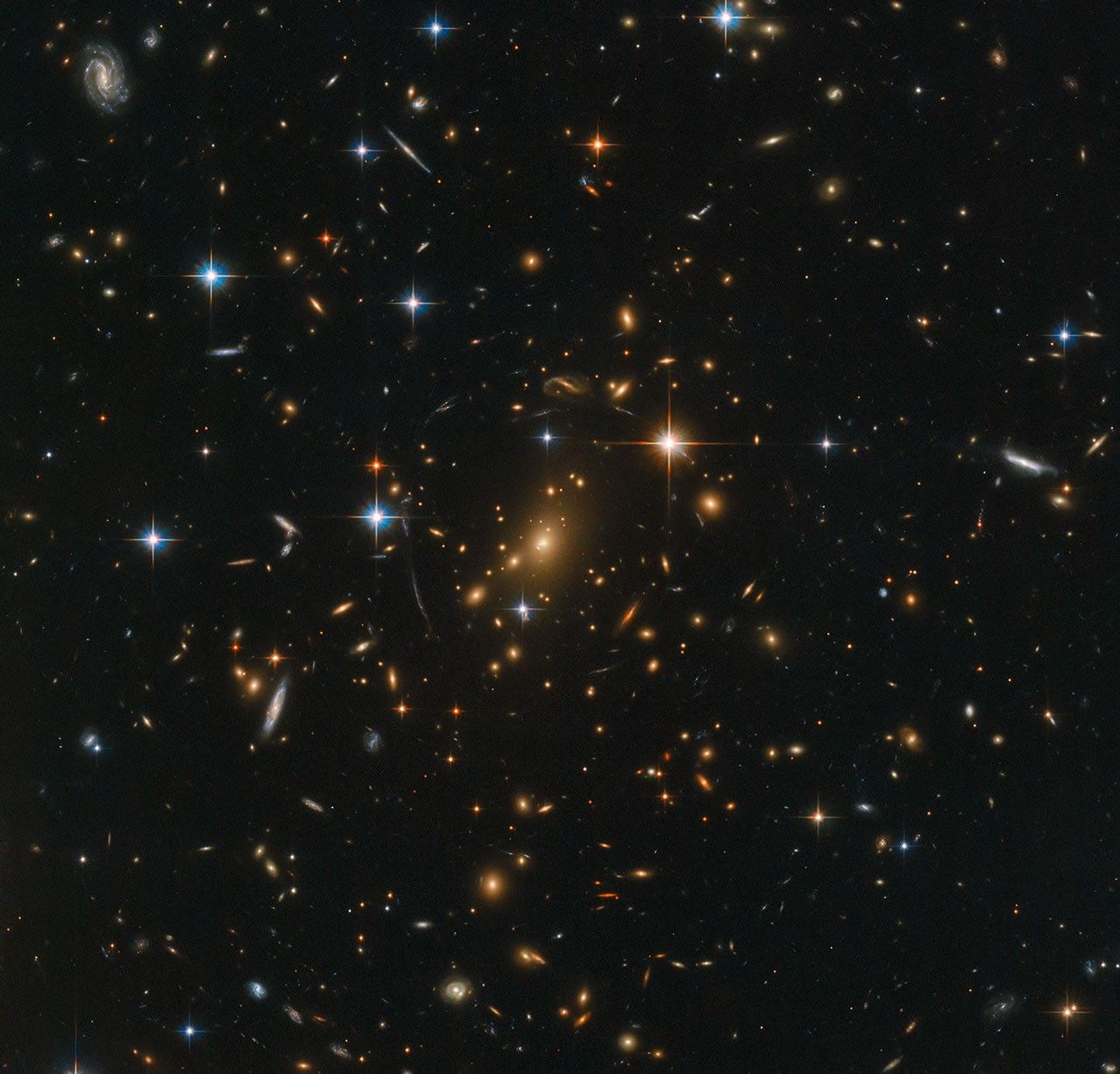 A packed field of galaxies and with faint curved arcs. Large grouping of galaxies warped around what appears to be a giant lens. Each galaxy appears as a glob of light, most of them reddish-orange. Bright twinkling foreground stars are scattered throughout the image.