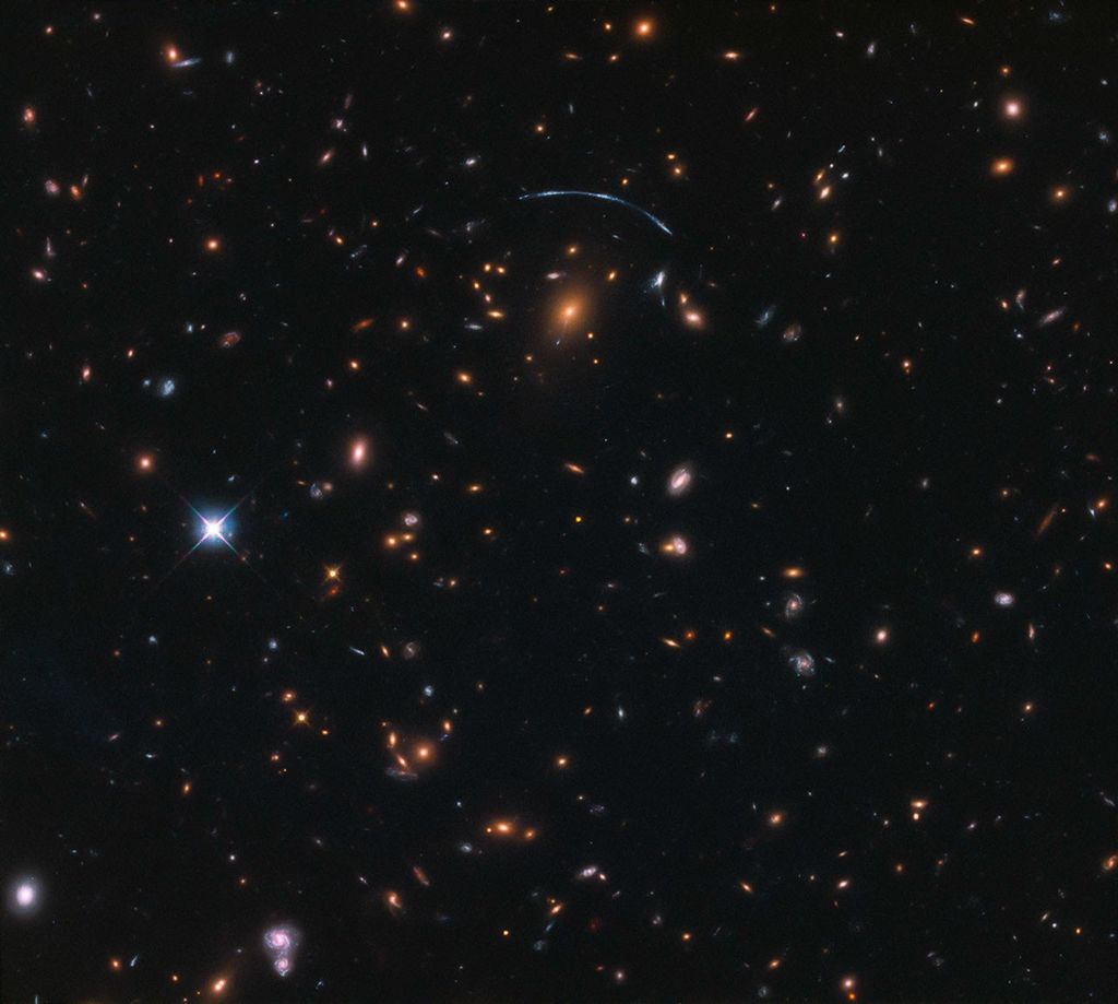 Black background dotted with galaxies. A reddish galaxy at image center with a bluish-white arc above it.