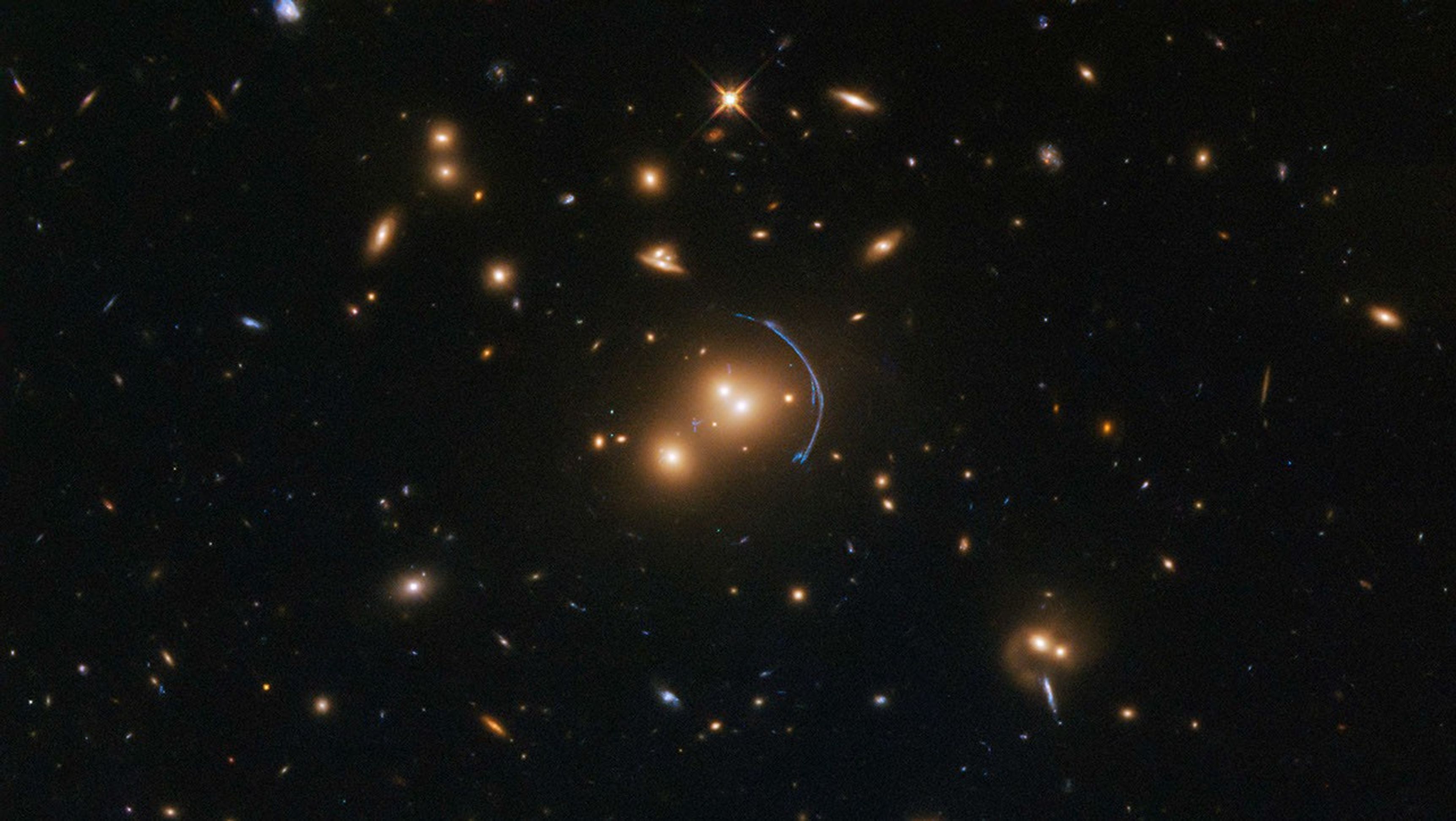 A field full of distant galaxies on a dark background. Most of them are red-orange spirals or spheres, but some are other colors and/or shapes. In the very center is a quartet of reddish elliptical galaxies, in two pairs of two dots close together. A blue-white, warped streak of light arcs around them on the right.