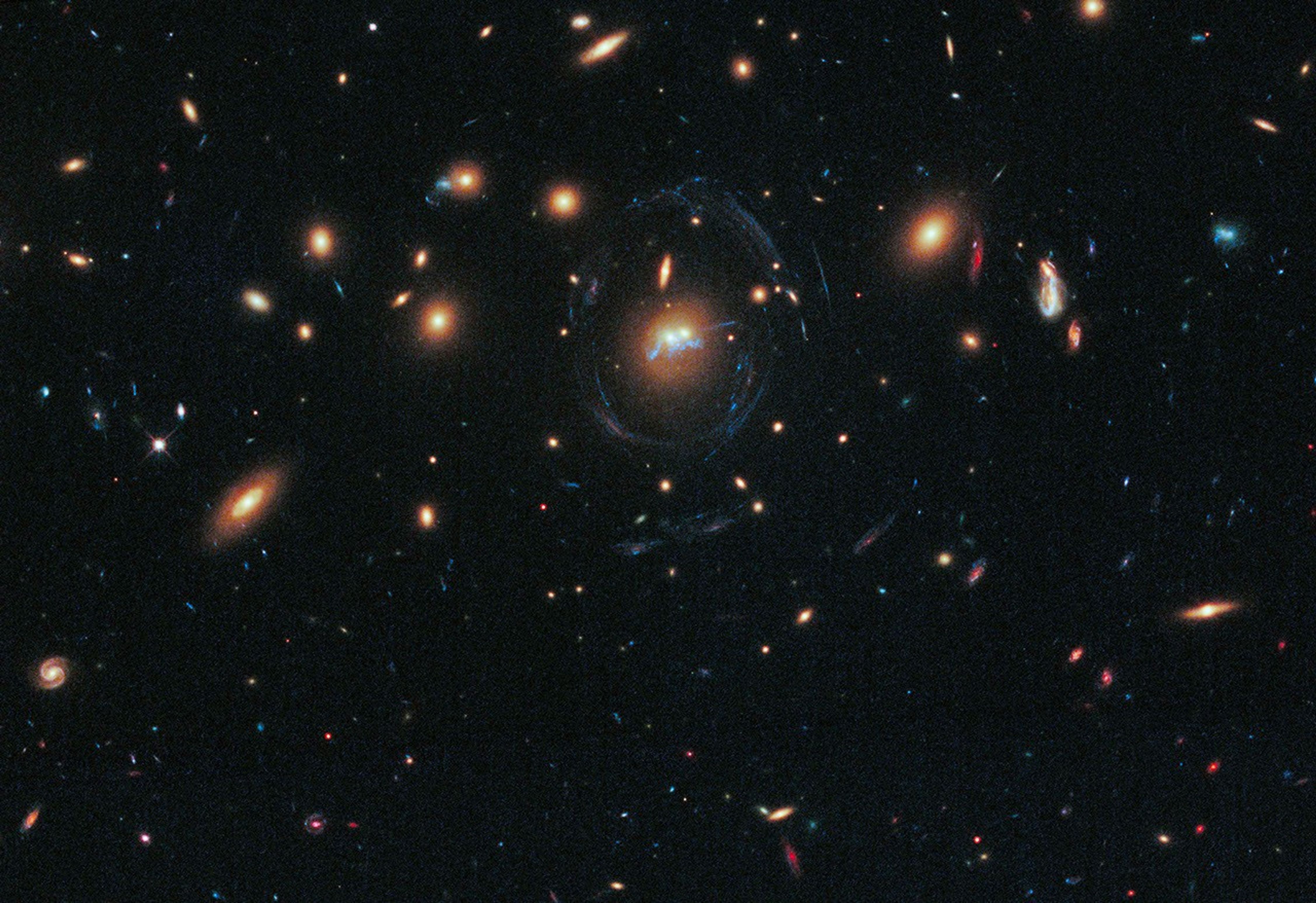 Black background dotted with galaxies. Two reddish galaxies at image center as two dots next to each other, with a thin bluish-white ring curving almost completely around it. Directly below the two galaxies, the filaments appear as a squiggly blue line.