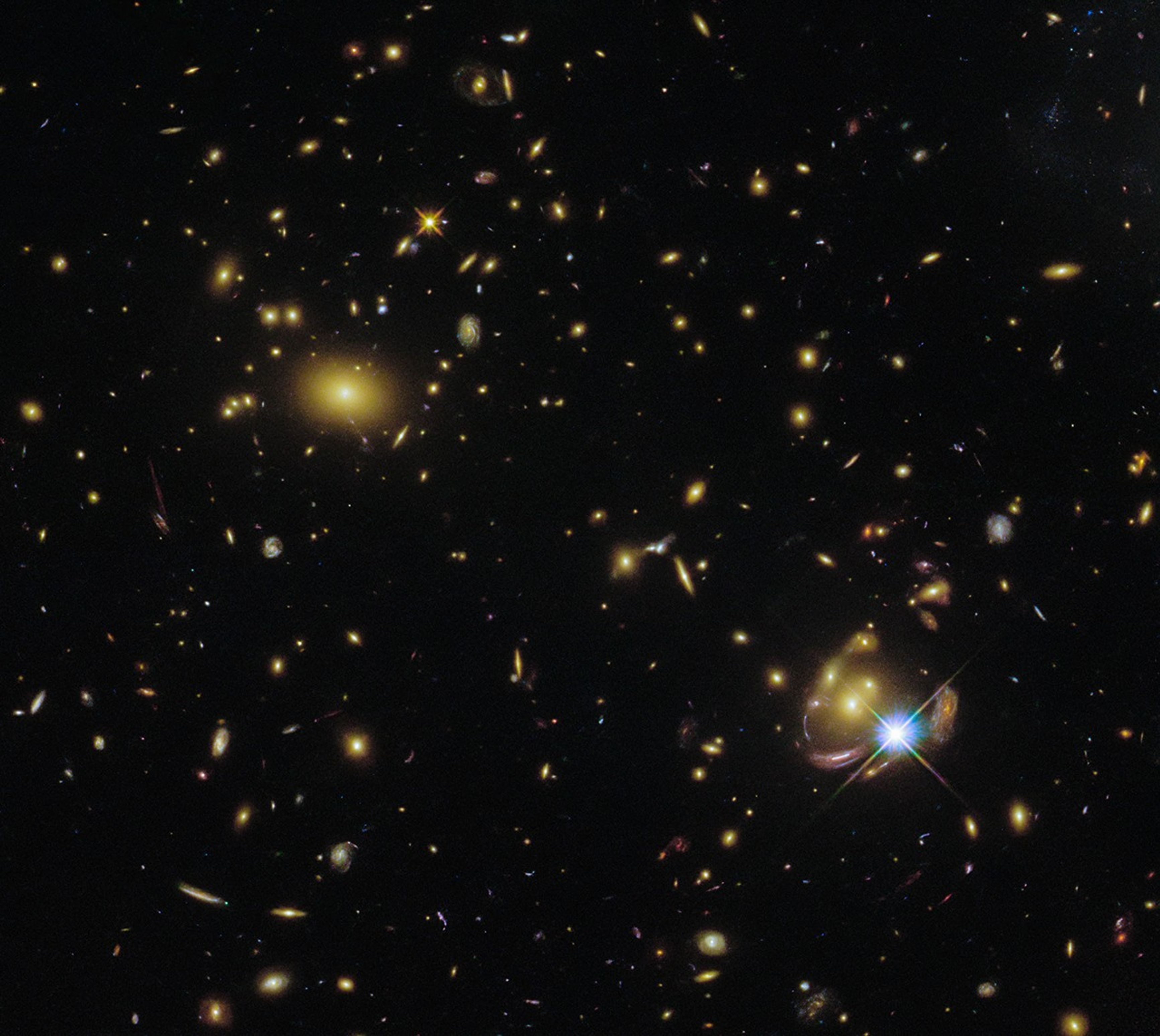 A cluster of various types of galaxies, visible as oval shapes in clumps. Two spiral galaxies are especially prominent near the center, and another on the right, where the lensing effect is best visible as a reddish curve with a bright star next to it and the same distorted galaxy appearing several times.