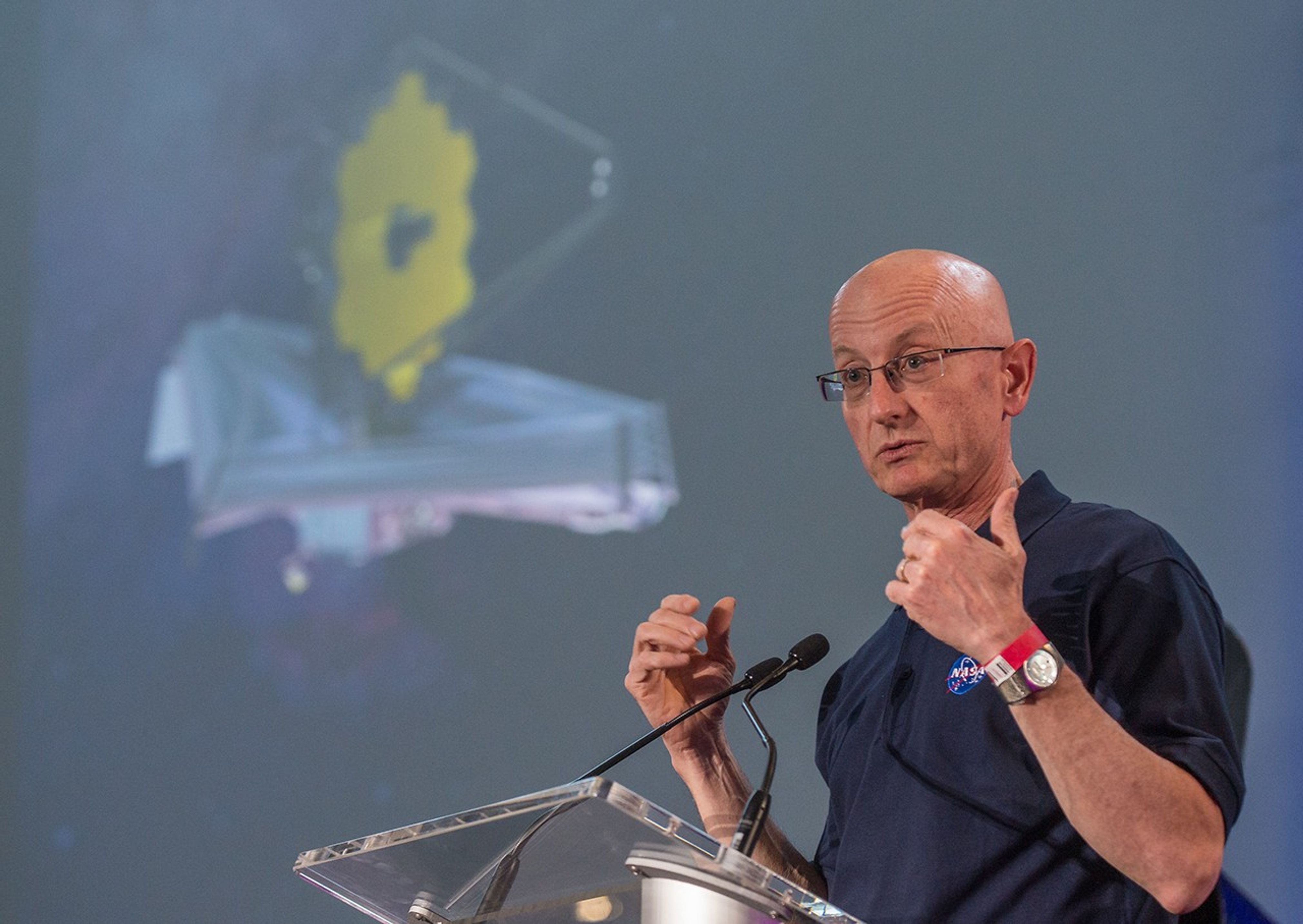 Eric Smith stands at the podium, addressing the audience, while an image of the James Webb Space Telescope is on the screen in the background.