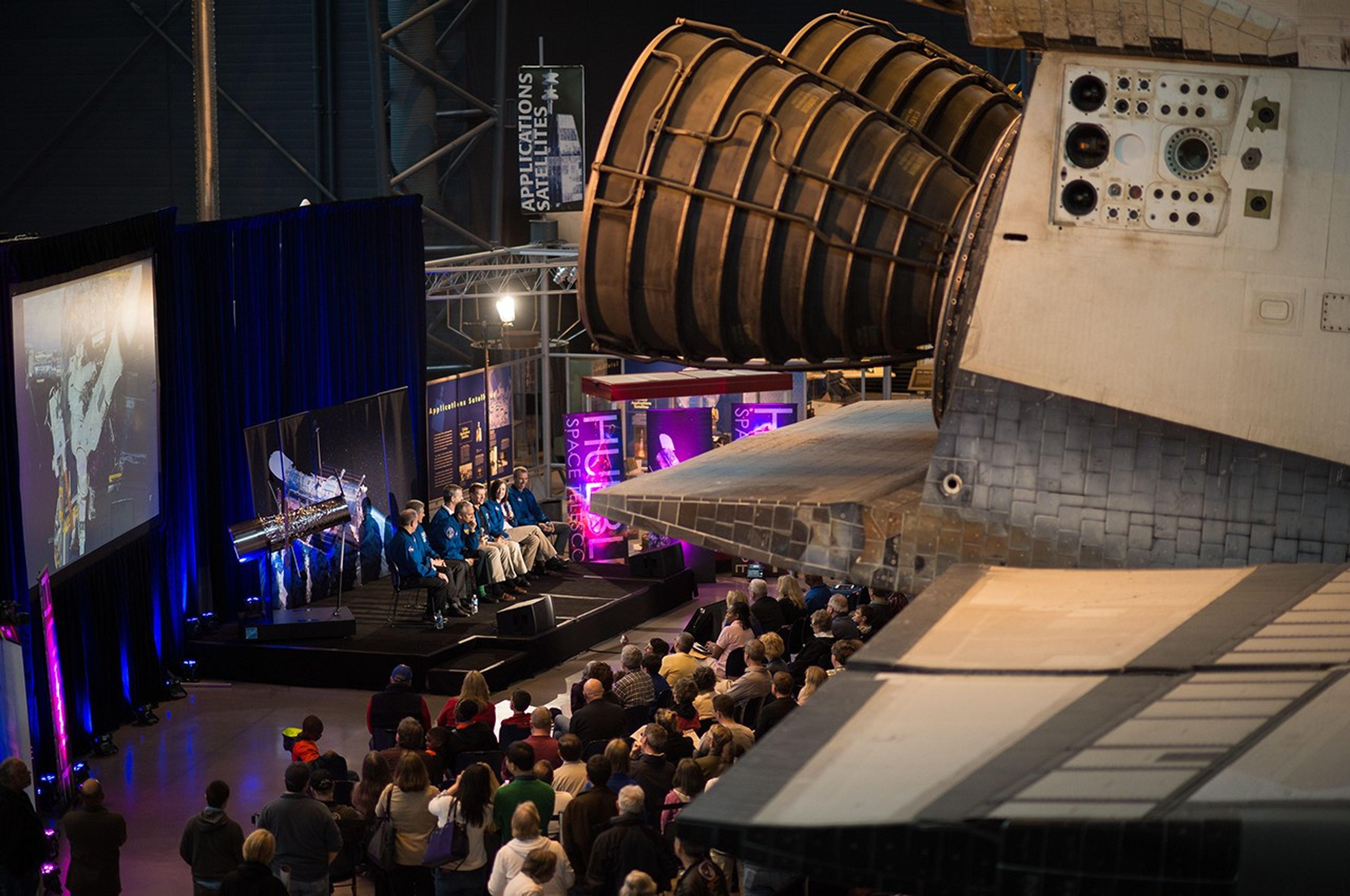 A photo taken from above and from a distance. The right half of the picture is mostly taken up by the back of Space Shuttle Discovery in the foreground. On the left, the astronauts sit on stage for the panel, with a model of Hubble behind them to the left, and a large audience listening.
